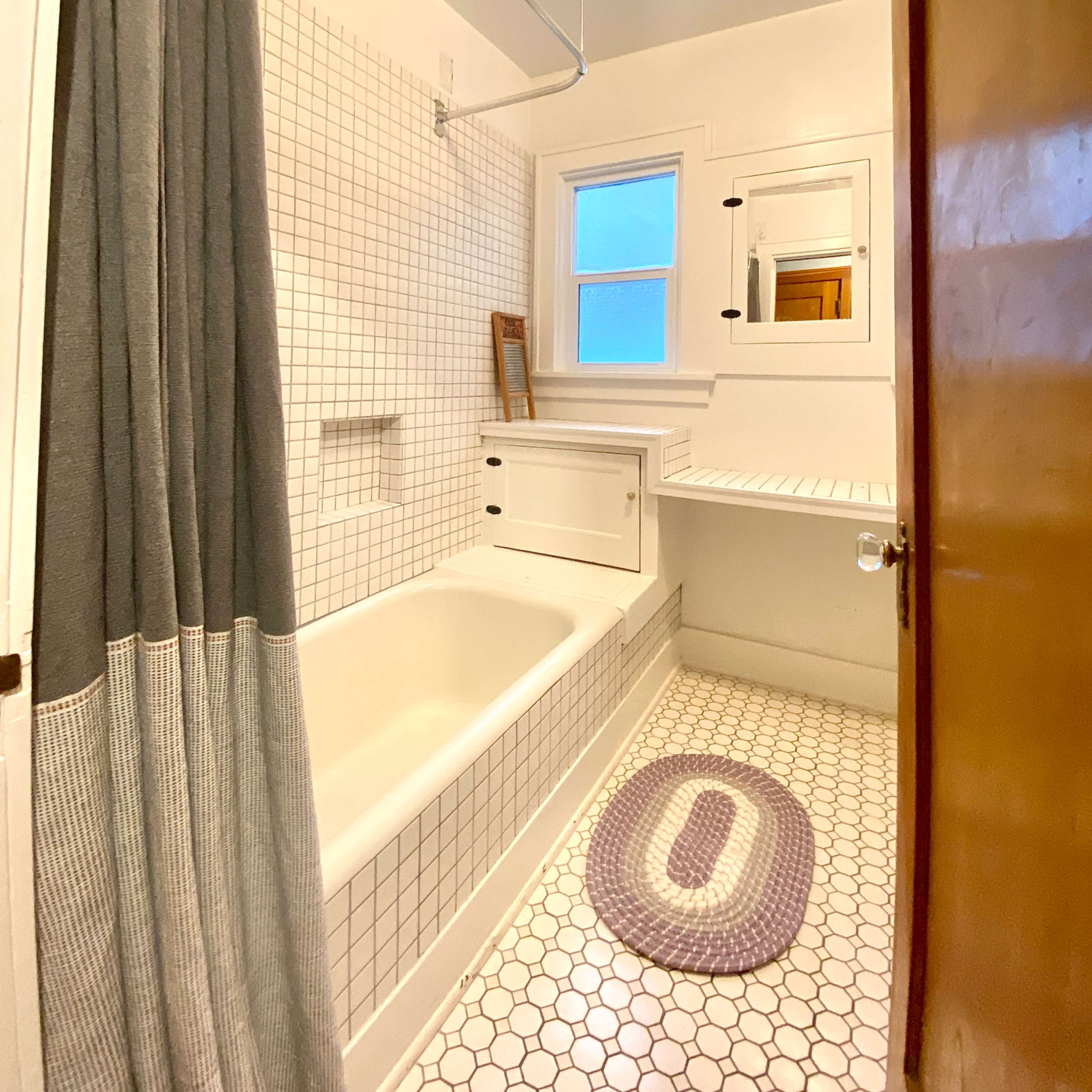A small bathroom features a white tiled bathtub, a gray shower curtain, a wall-mounted mirror, and a circular rug on the tile floor.