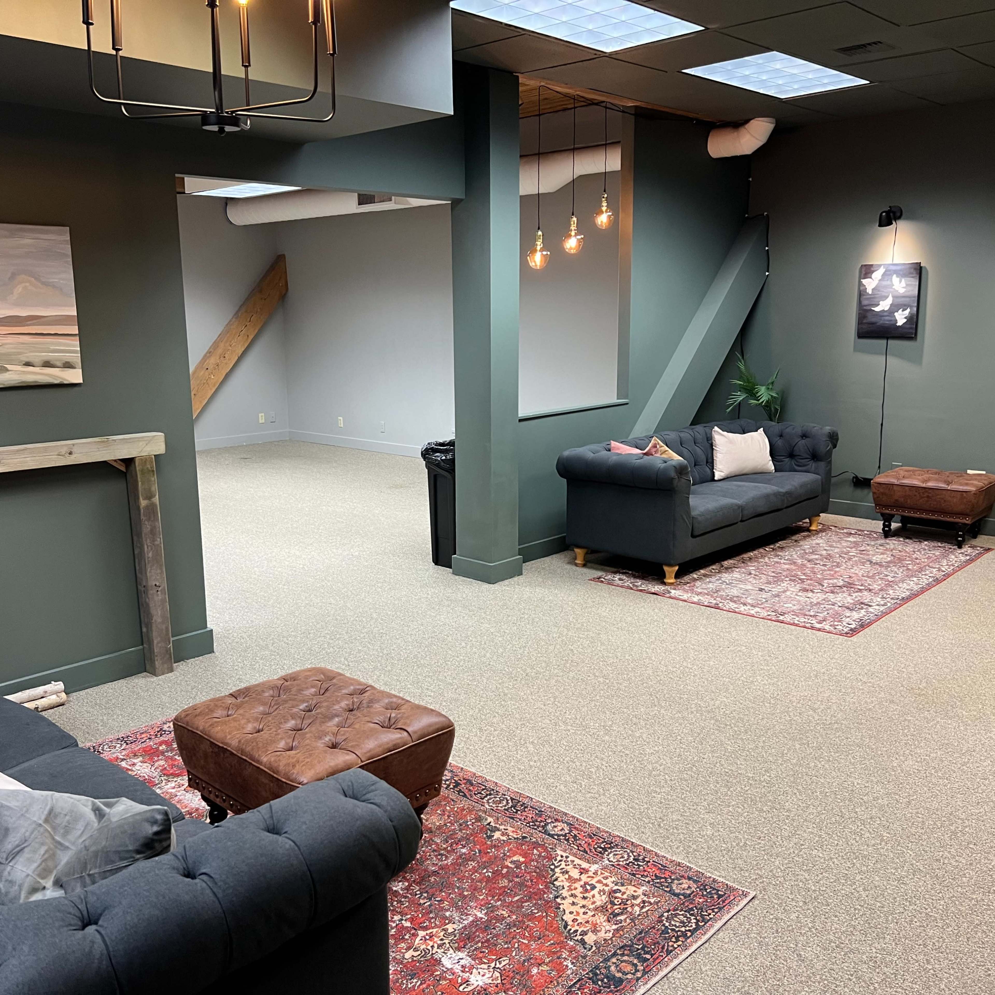A spacious, well-lit room featuring two dark sofas, a wooden table, and an area rug, with an abstract painting and decorative lighting on the walls.