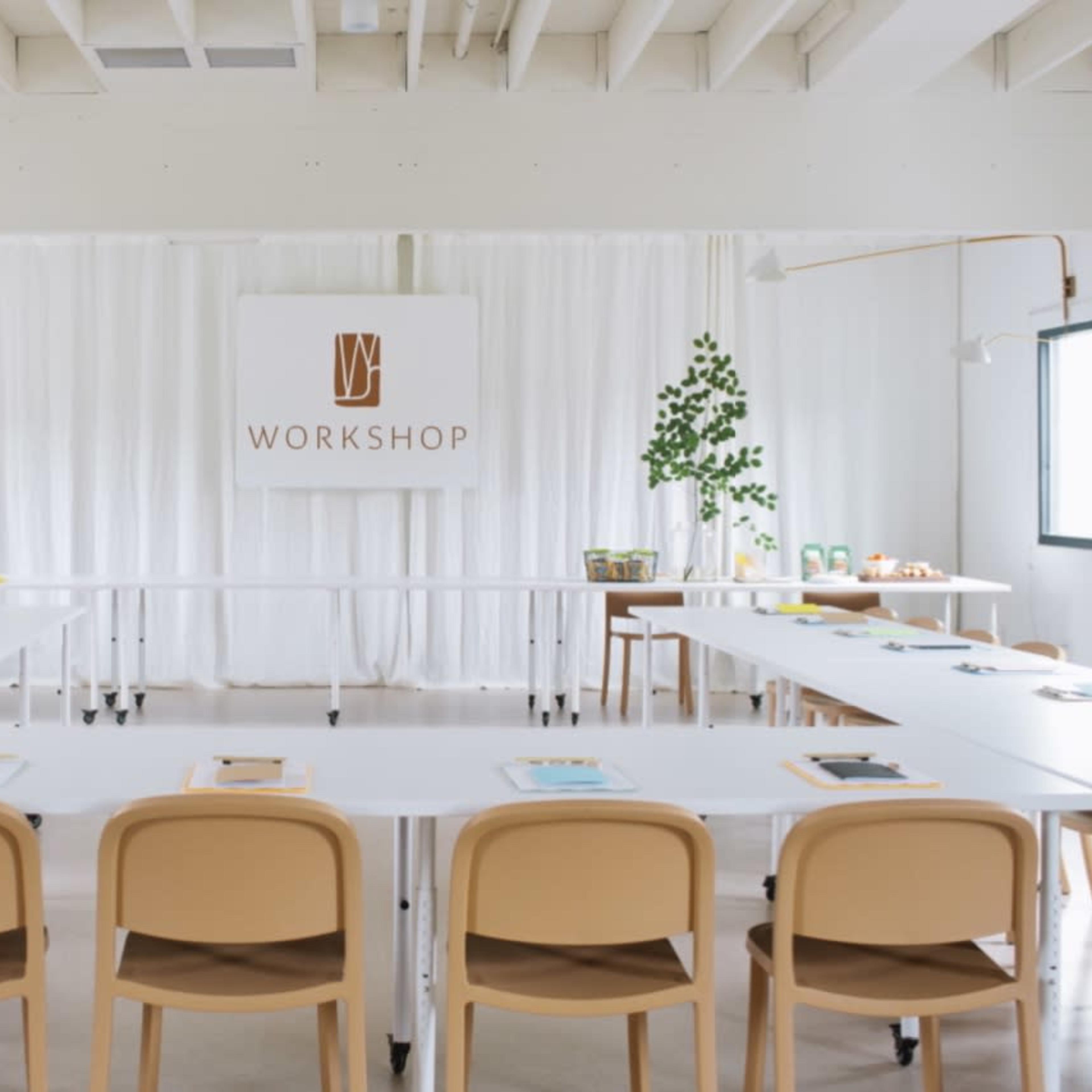A bright, modern workshop space features a long table set with materials, surrounded by light-colored chairs and green plants.