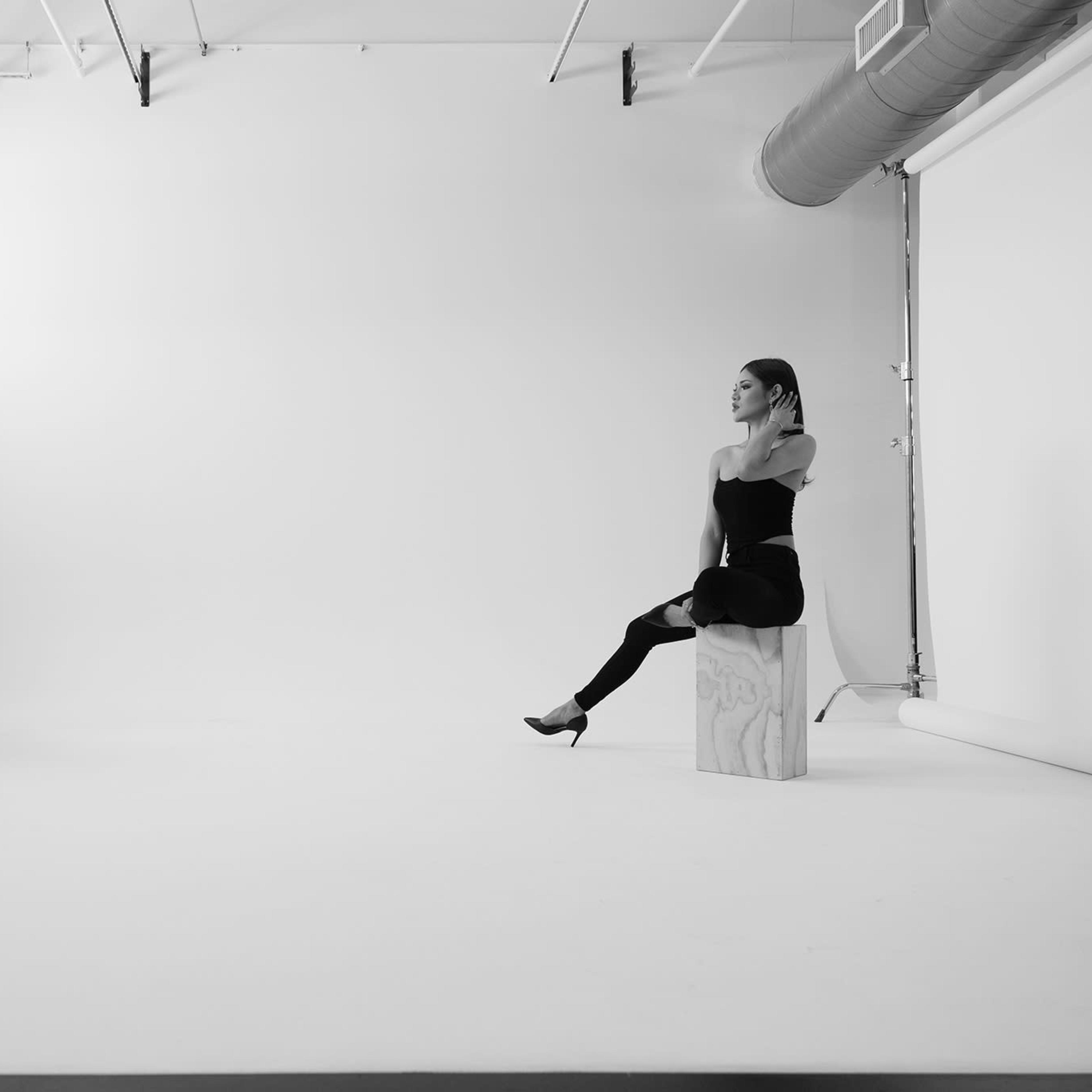 A photographer is taking pictures of a model seated on a wooden block in a stark, empty studio.