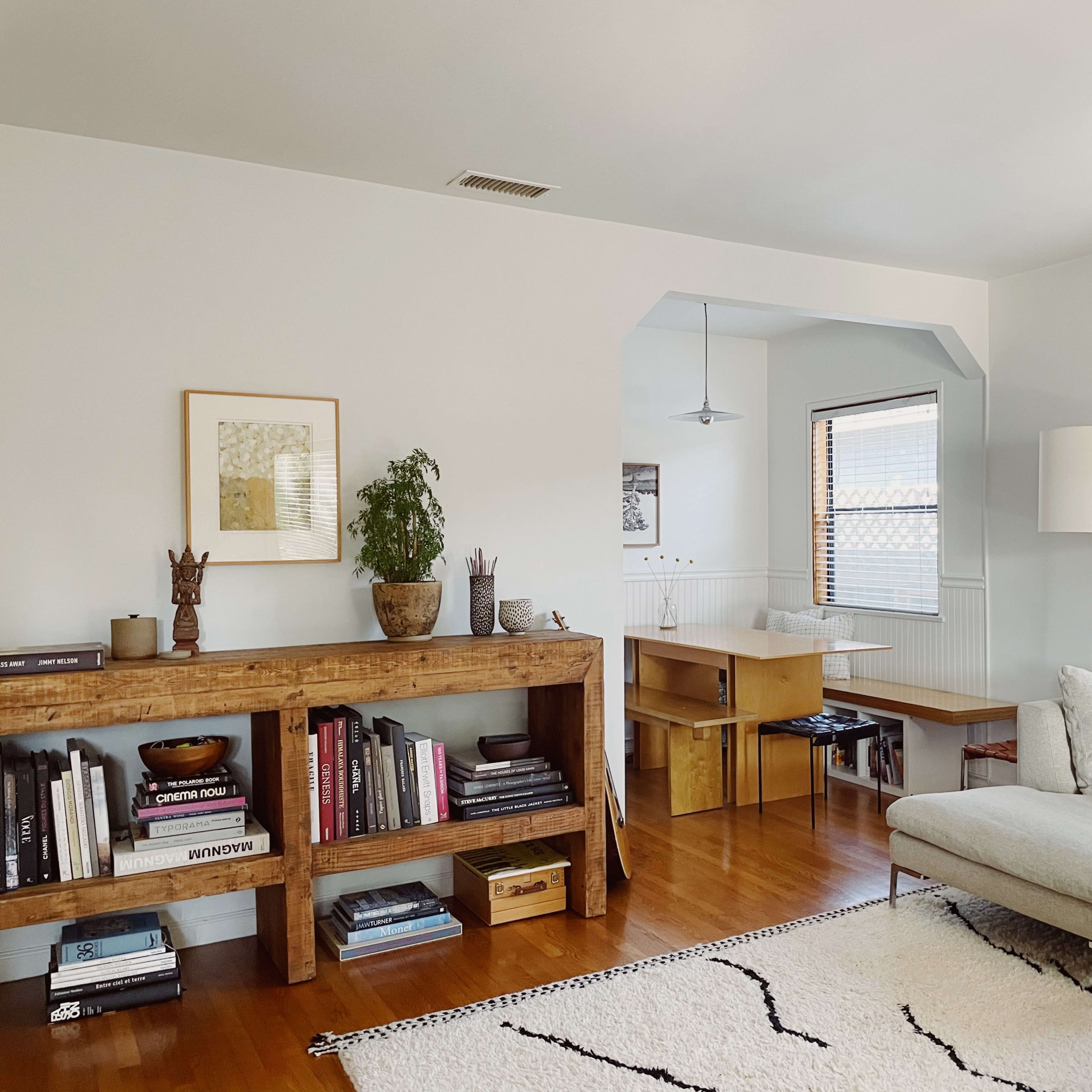 A modern living room with a wooden bookshelf, a light gray sectional sofa, and a small dining area in the background.