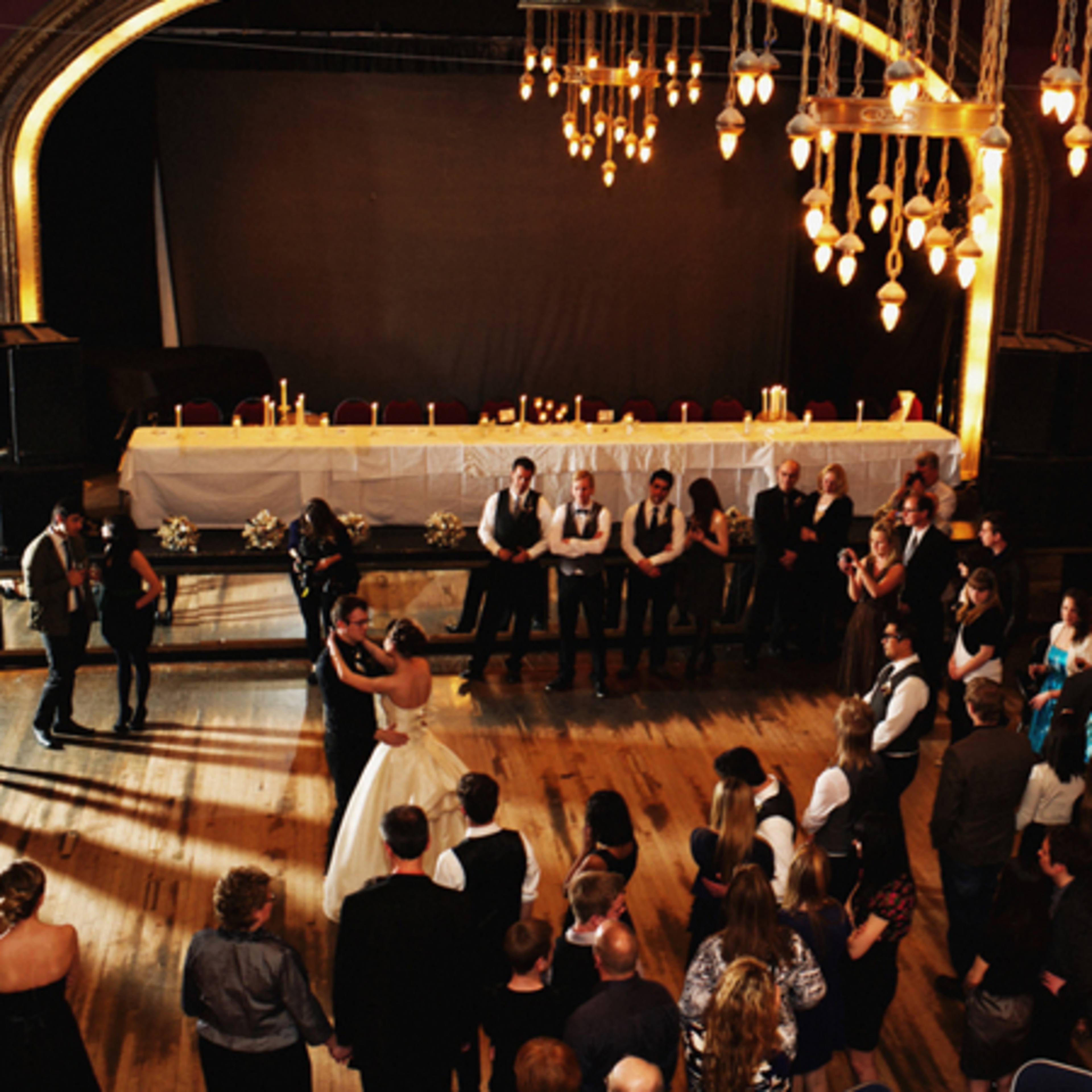 A bride and groom dance in the center of a decorated venue surrounded by guests seated at tables.