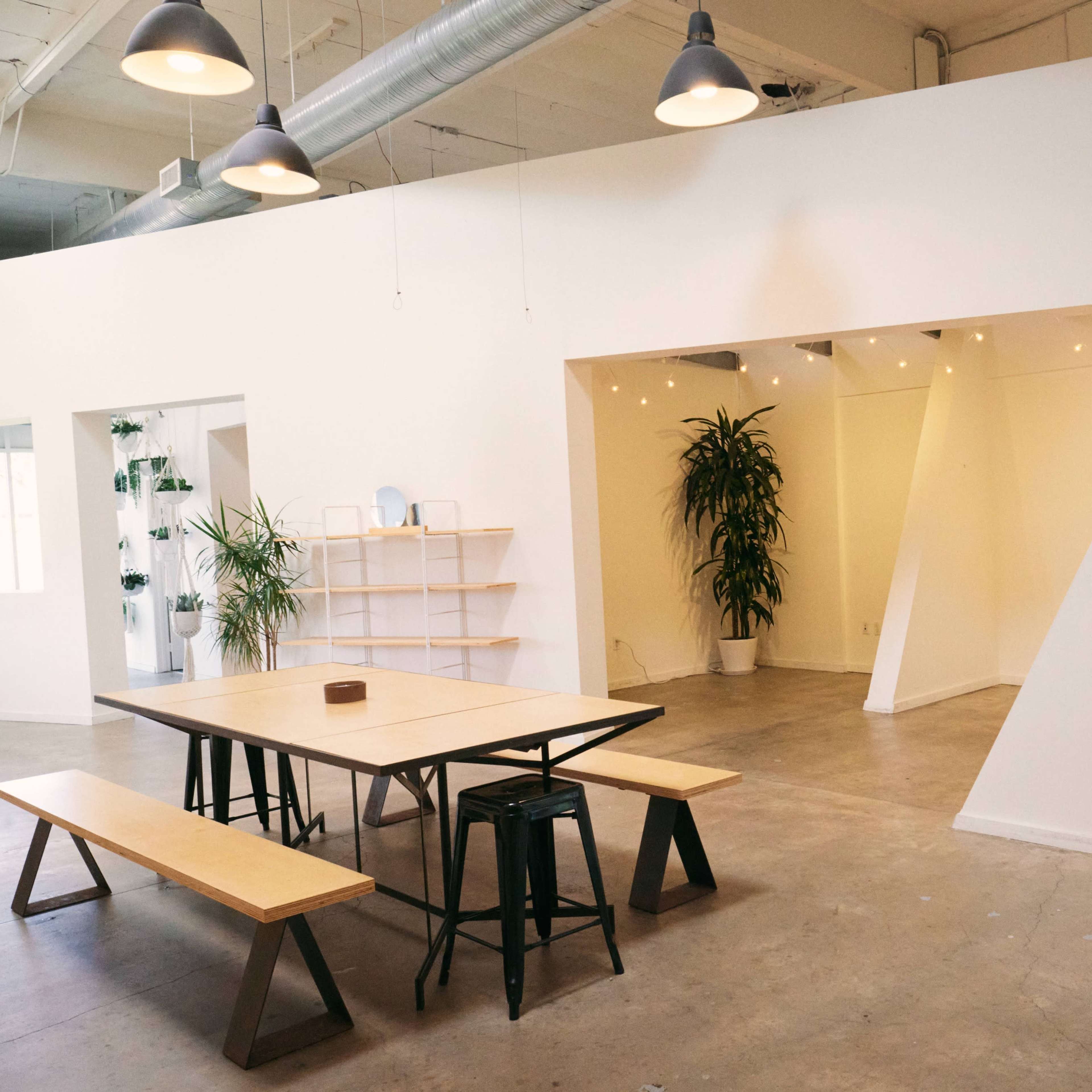 A modern, minimalist interior space featuring a wooden table with benches, surrounded by potted plants and white walls.