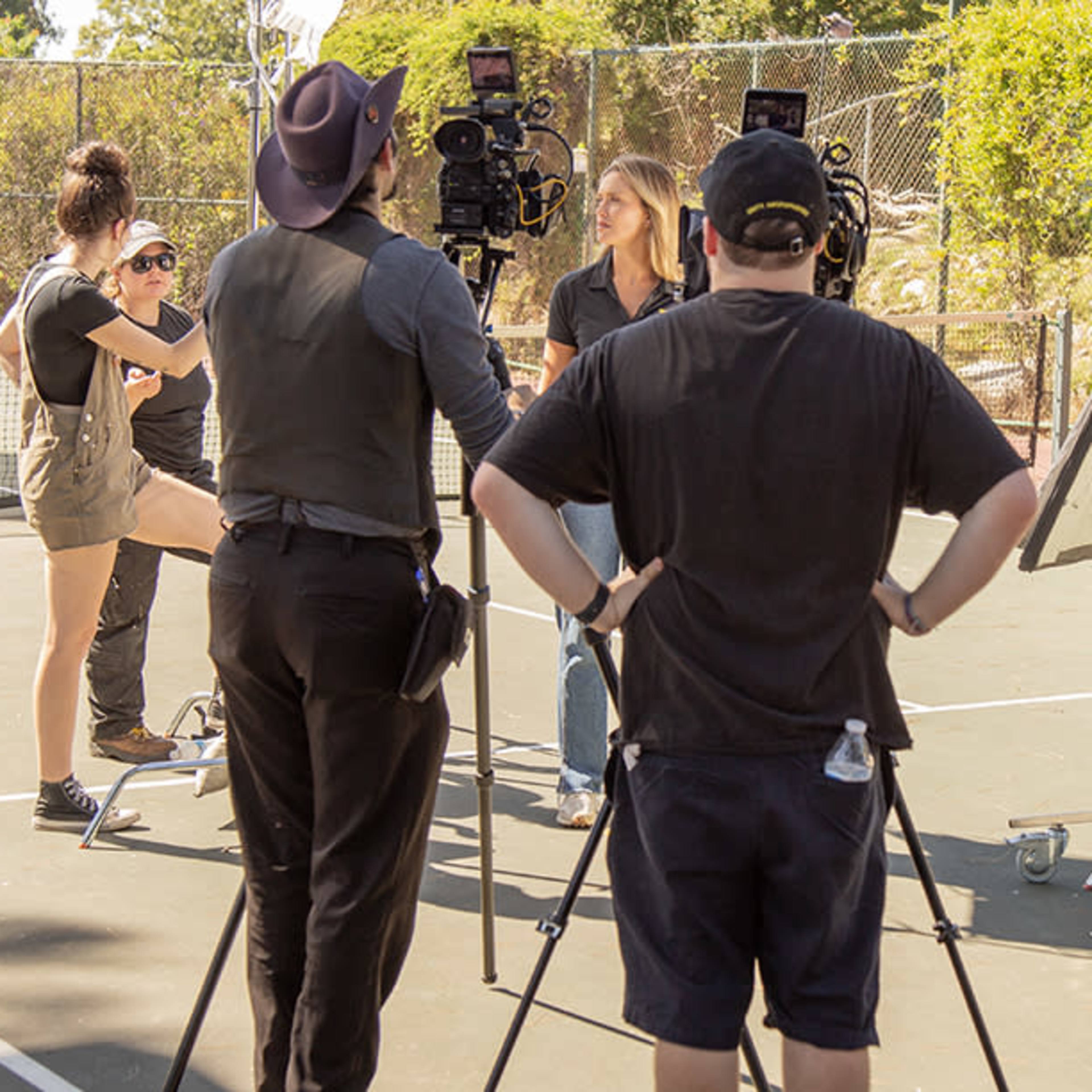 A film crew sets up on a tennis court, with cameras and equipment directed towards a woman who is being interviewed.
