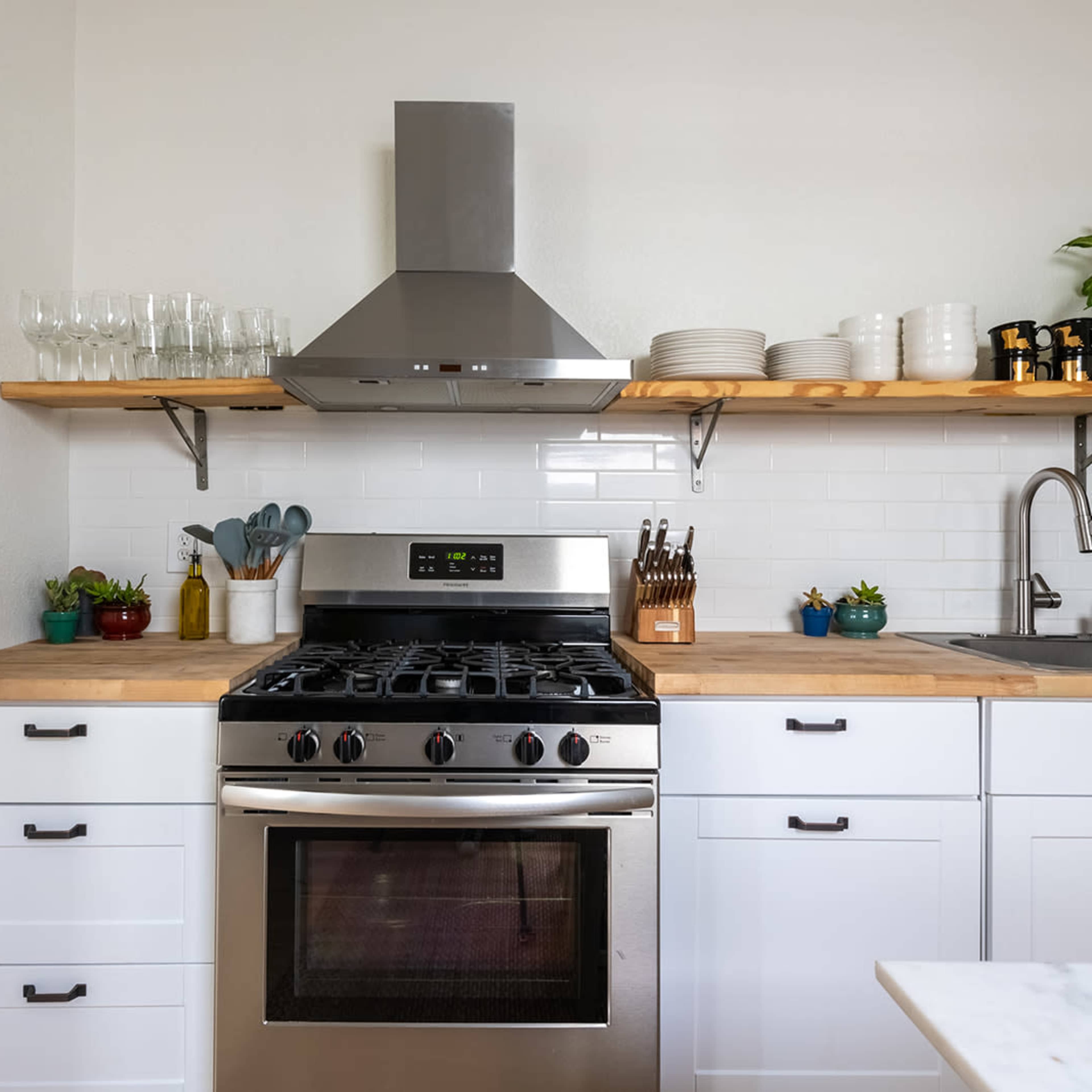 A modern kitchen featuring a stainless steel range hood above a gas stove, white cabinetry, and wooden open shelves with various glassware and plants.