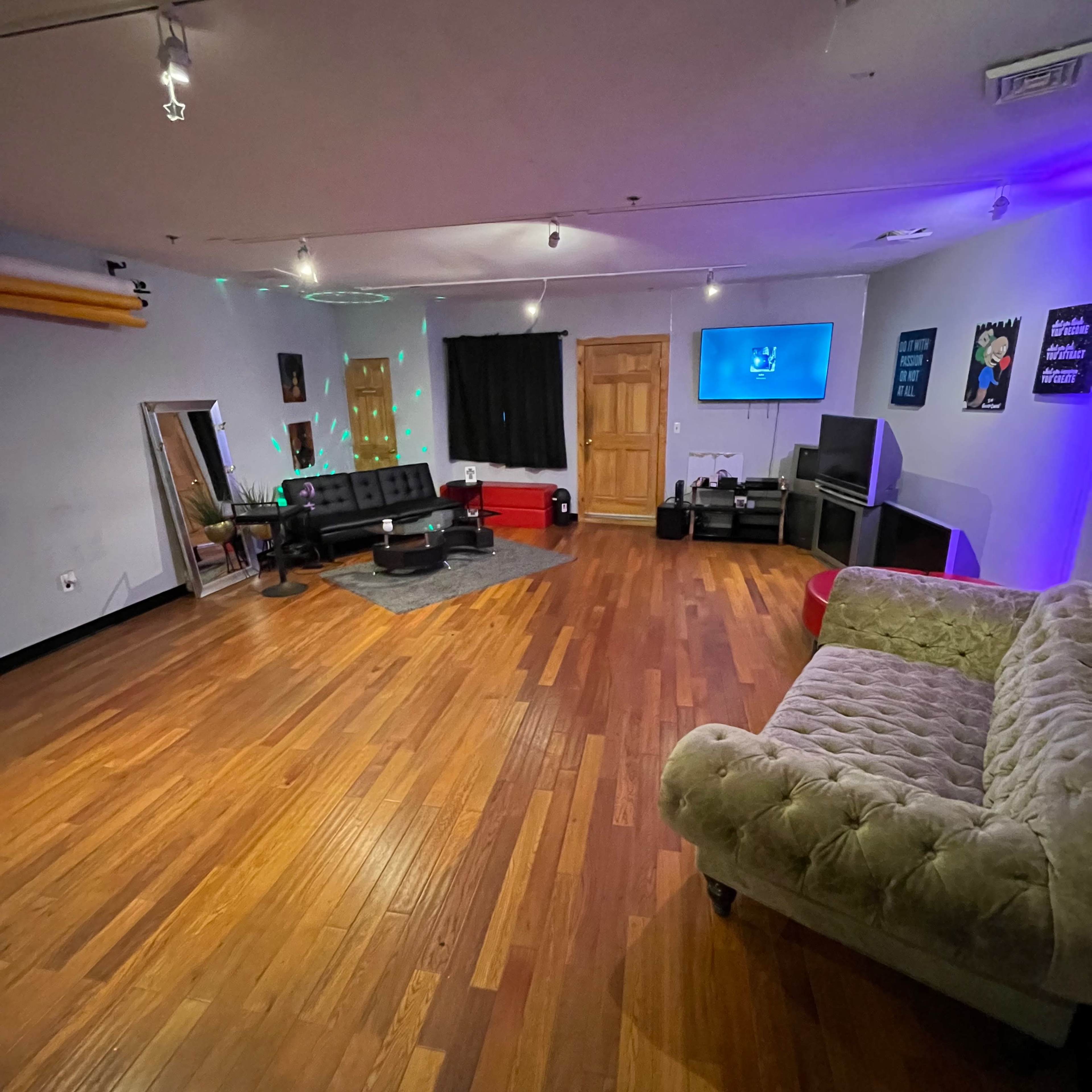 The room features hardwood flooring, a mix of seating arrangements, a wall-mounted TV, and ambient lighting with a black curtain on one side.