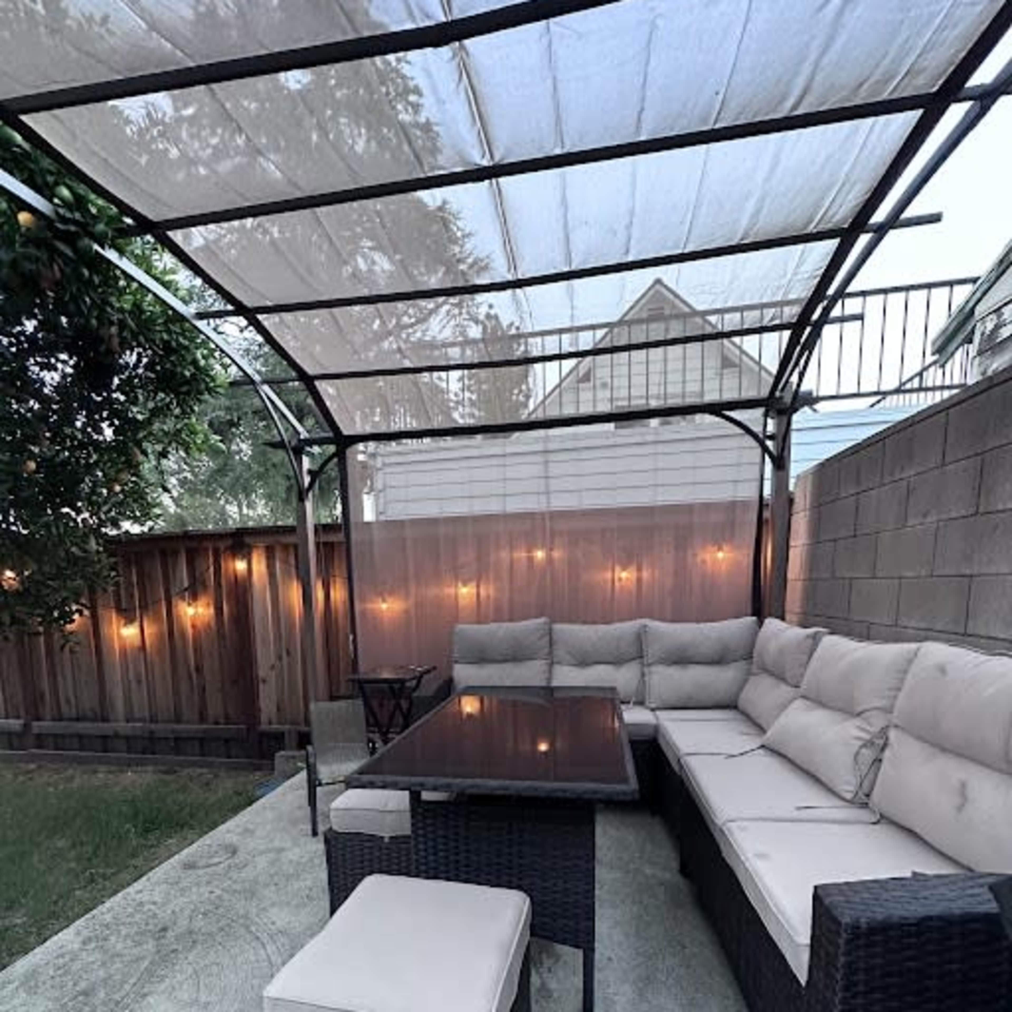 A covered outdoor seating area features a sectional sofa, a table, and additional seating against a backdrop of wooden fencing and soft lighting.