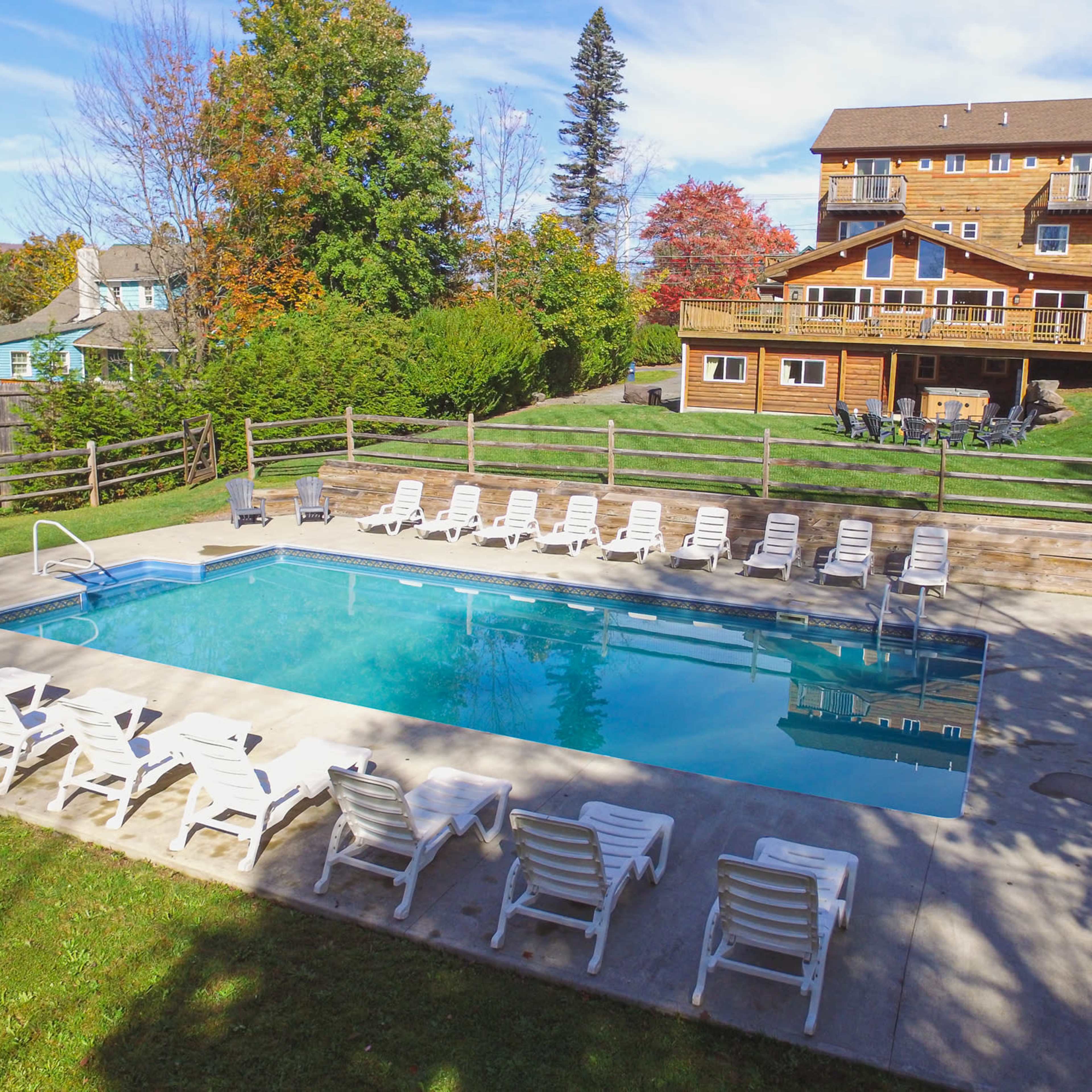 A large outdoor swimming pool is surrounded by lounge chairs, with a multi-story wooden building and trees in the background.