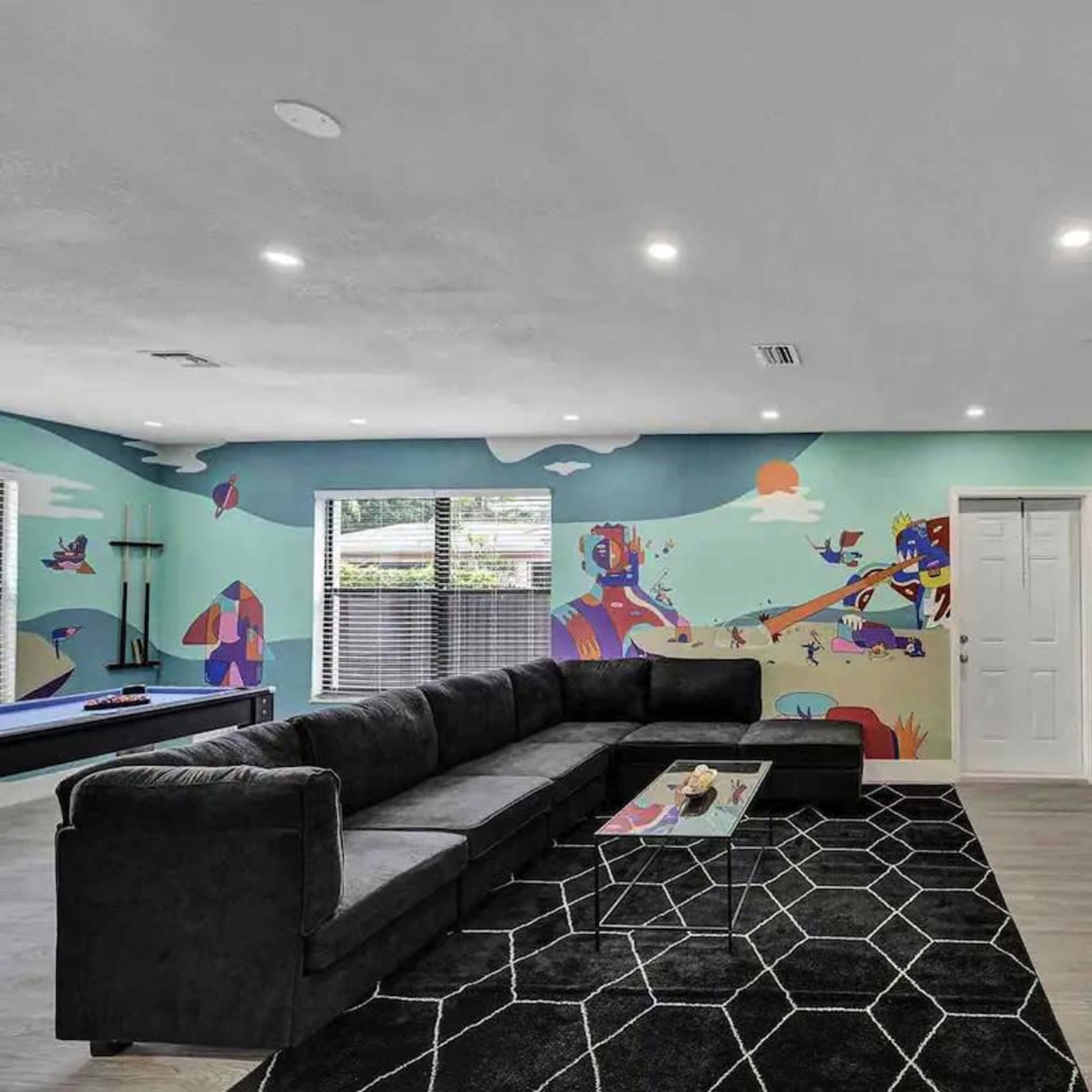 The living room features a large sectional sofa, a colorful mural on the wall, a TV mounted on a side wall, and a modern black rug on the floor.