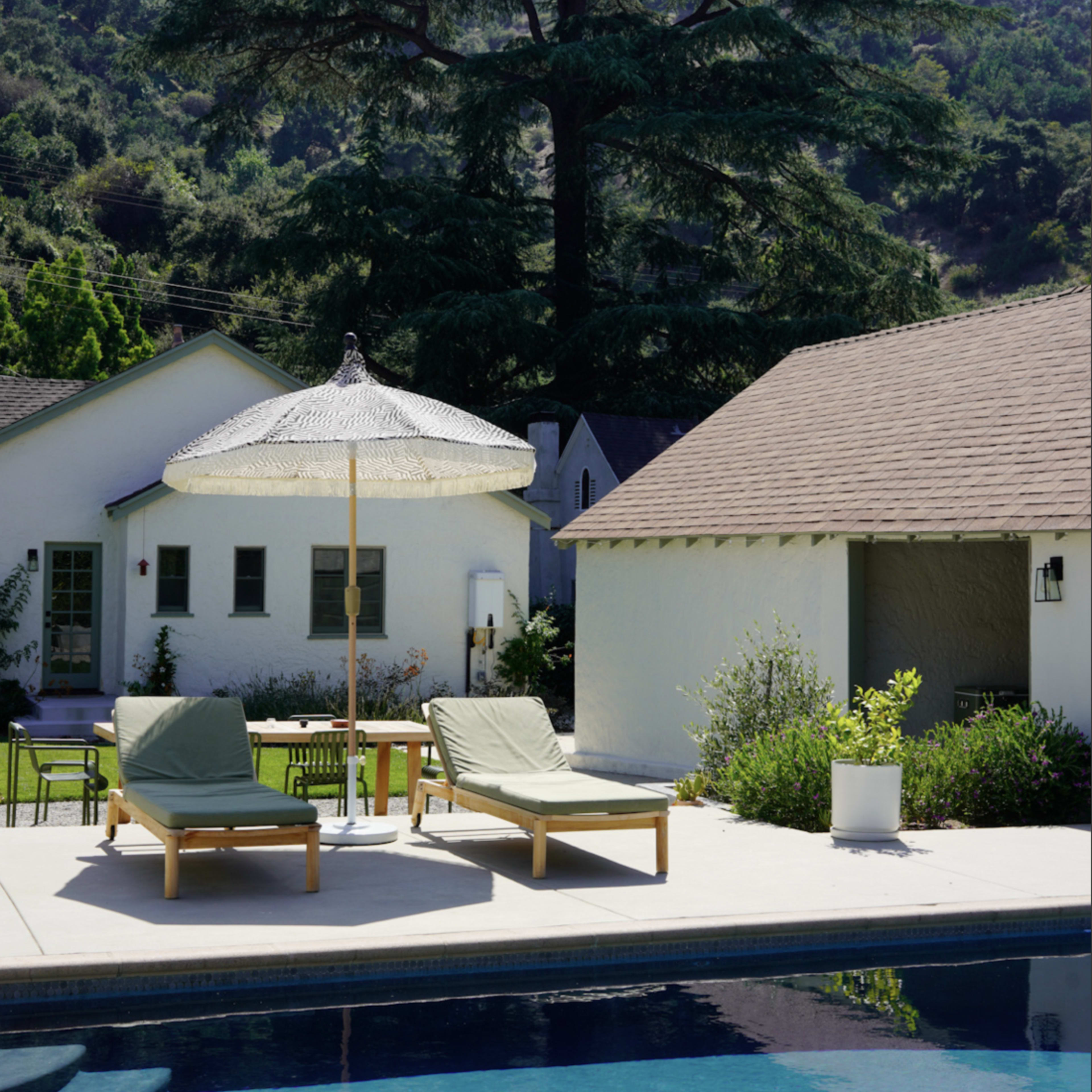A poolside area with lounge chairs, a large umbrella, and a backdrop of a tall tree and hillside.