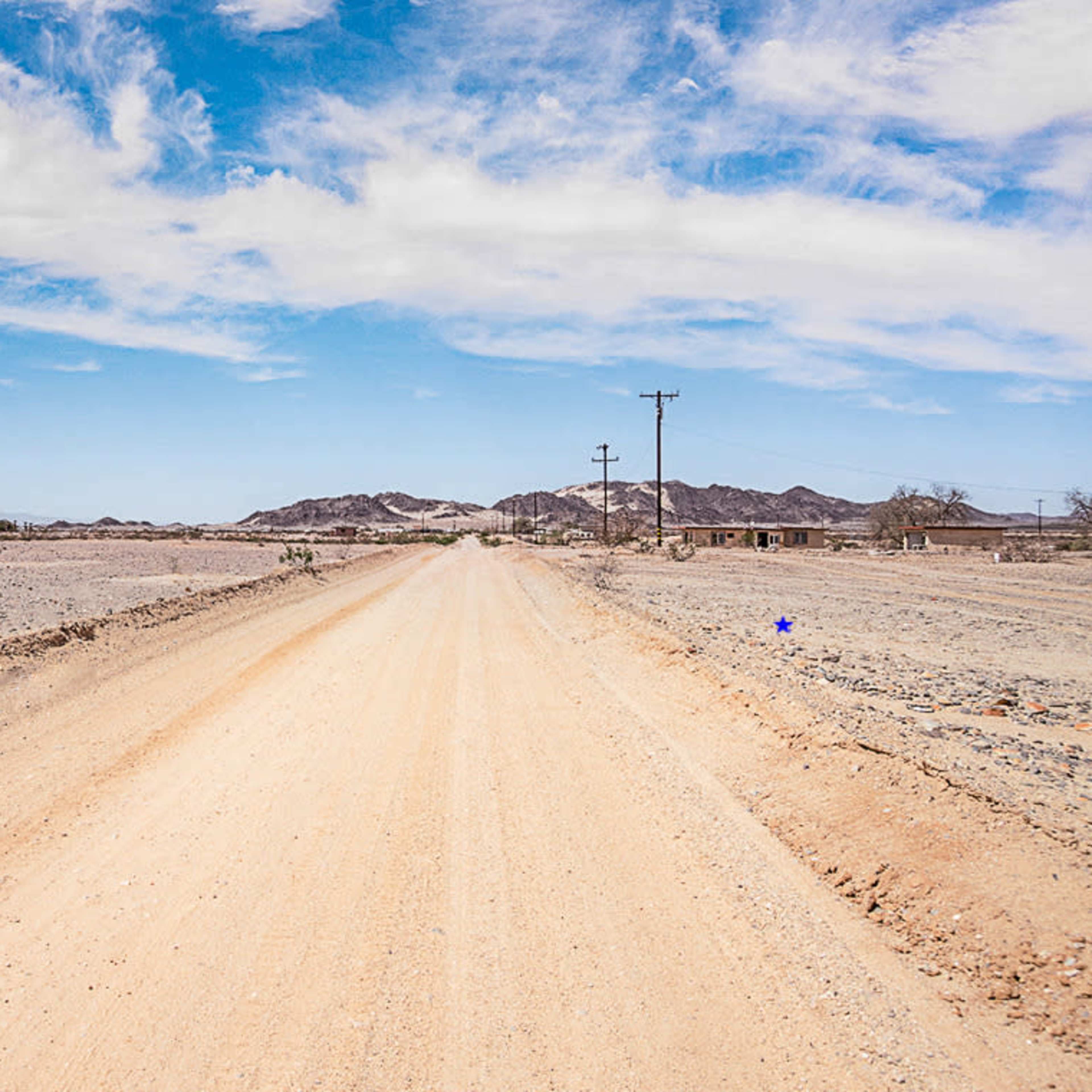 A dirt road stretches through a barren landscape, flanked by sparse vegetation and distant mountains under a blue sky.