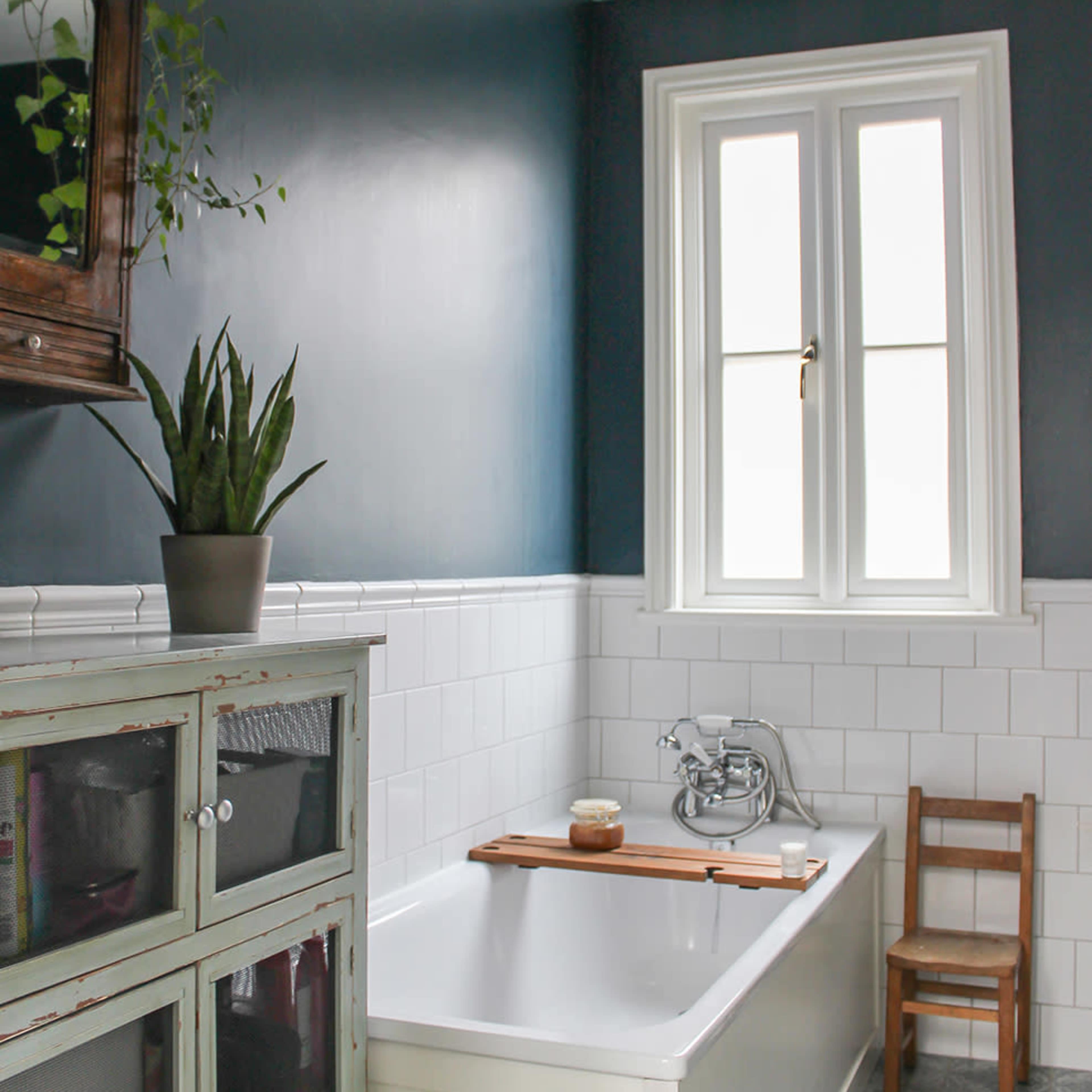 A modern bathroom featuring a freestanding bathtub, a small wooden chair, and a potted plant beside a vintage cabinet against a blue wall.
