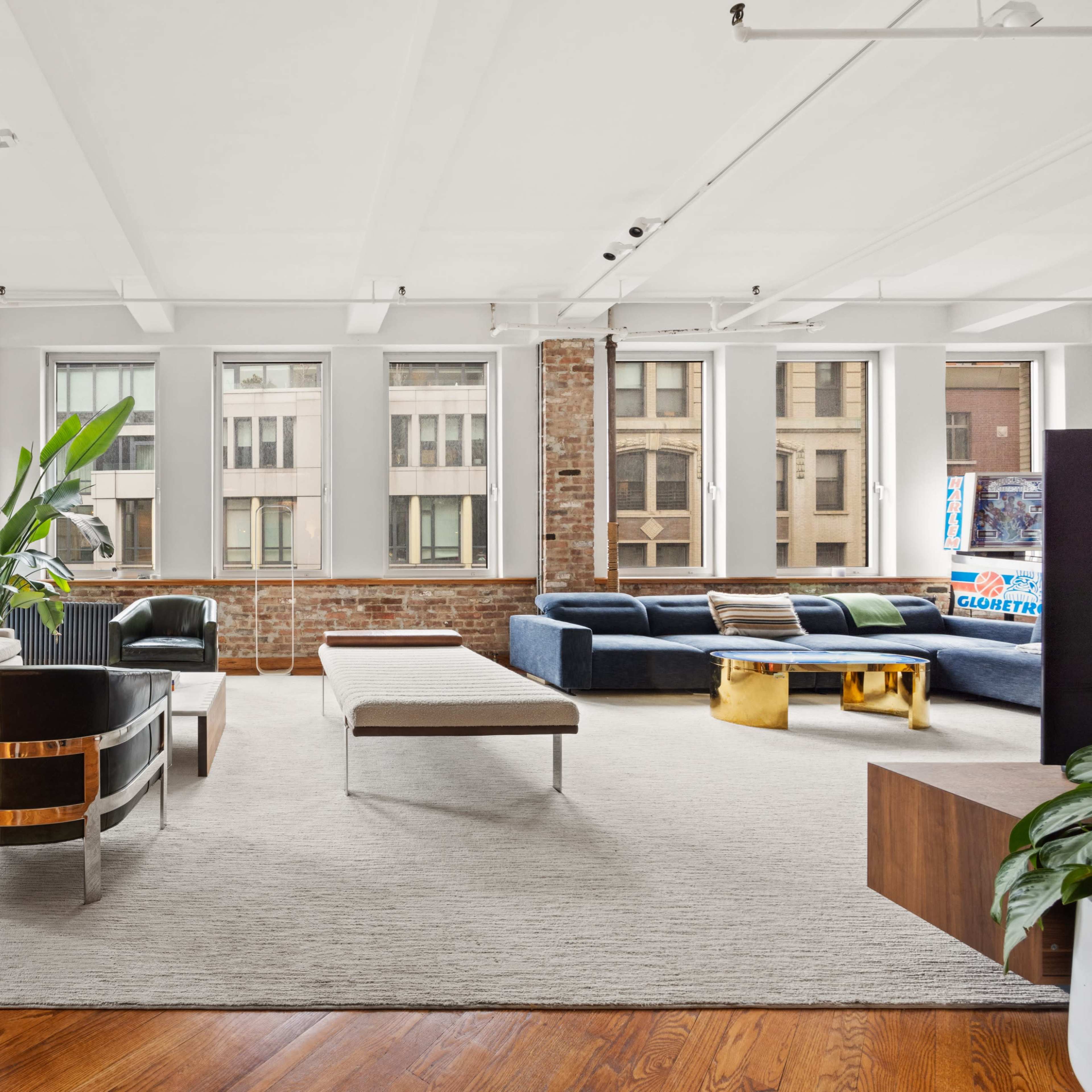 A spacious living room features large windows, exposed brick walls, a light-colored sofa, a dark blue sectional, and a stylish coffee table on a textured area rug.