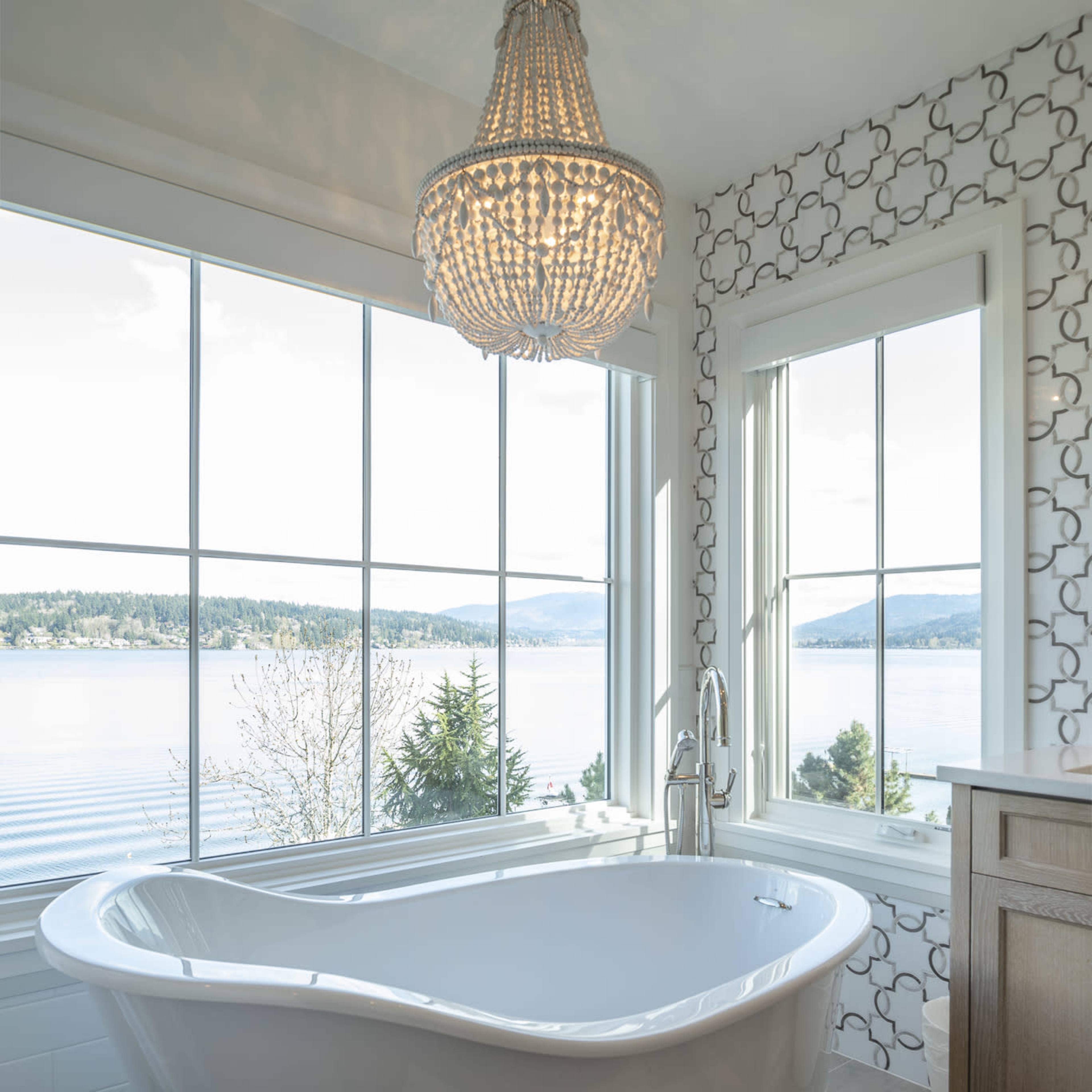 A freestanding white bathtub is positioned next to large windows, offering a view of a lake and trees while illuminated by a chandelier above.