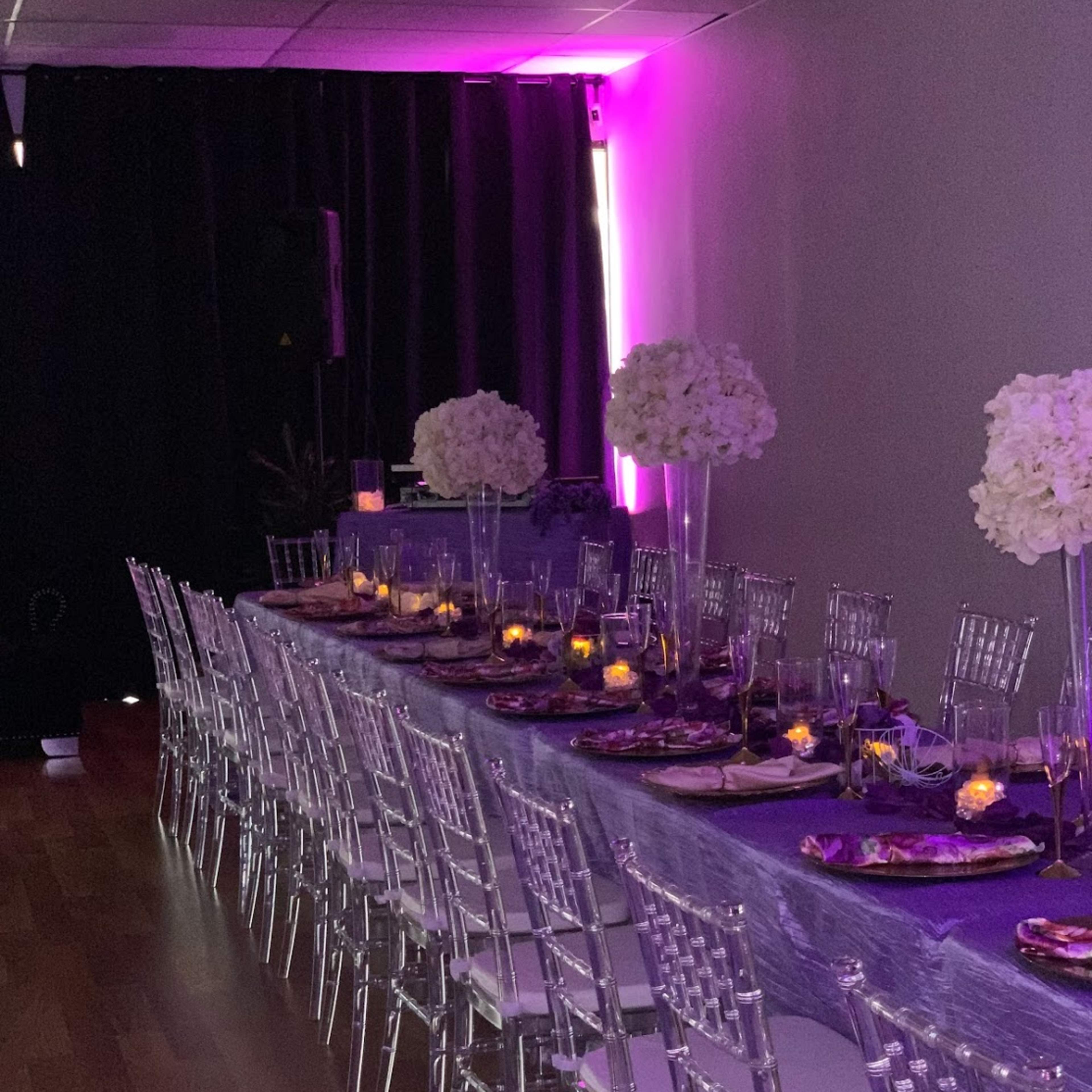 A long dining table is elegantly arranged with transparent chairs and centered floral arrangements, set against a backdrop of purple lighting.