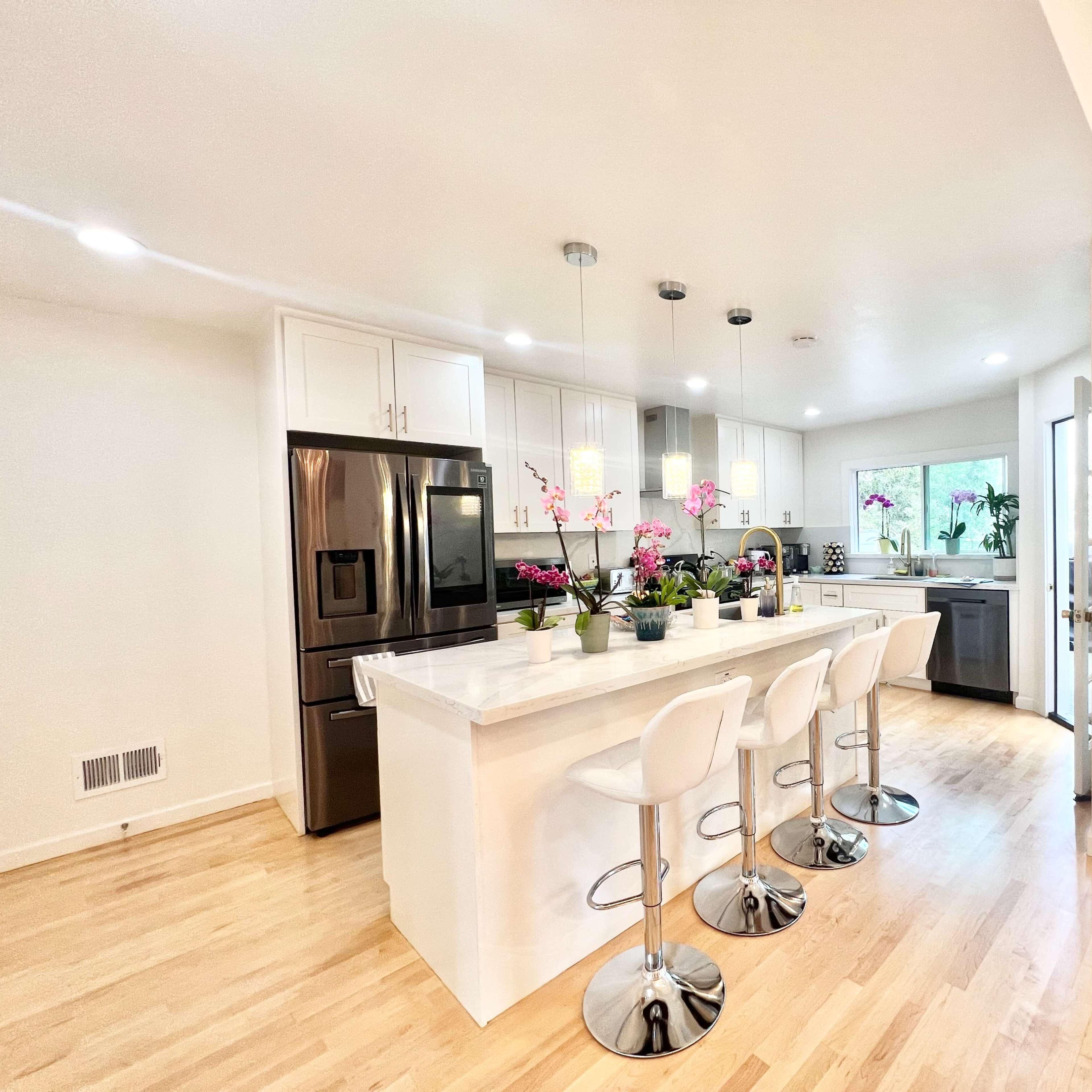 A modern kitchen features white cabinetry, a marble island with bar stools, and natural light coming through large windows.