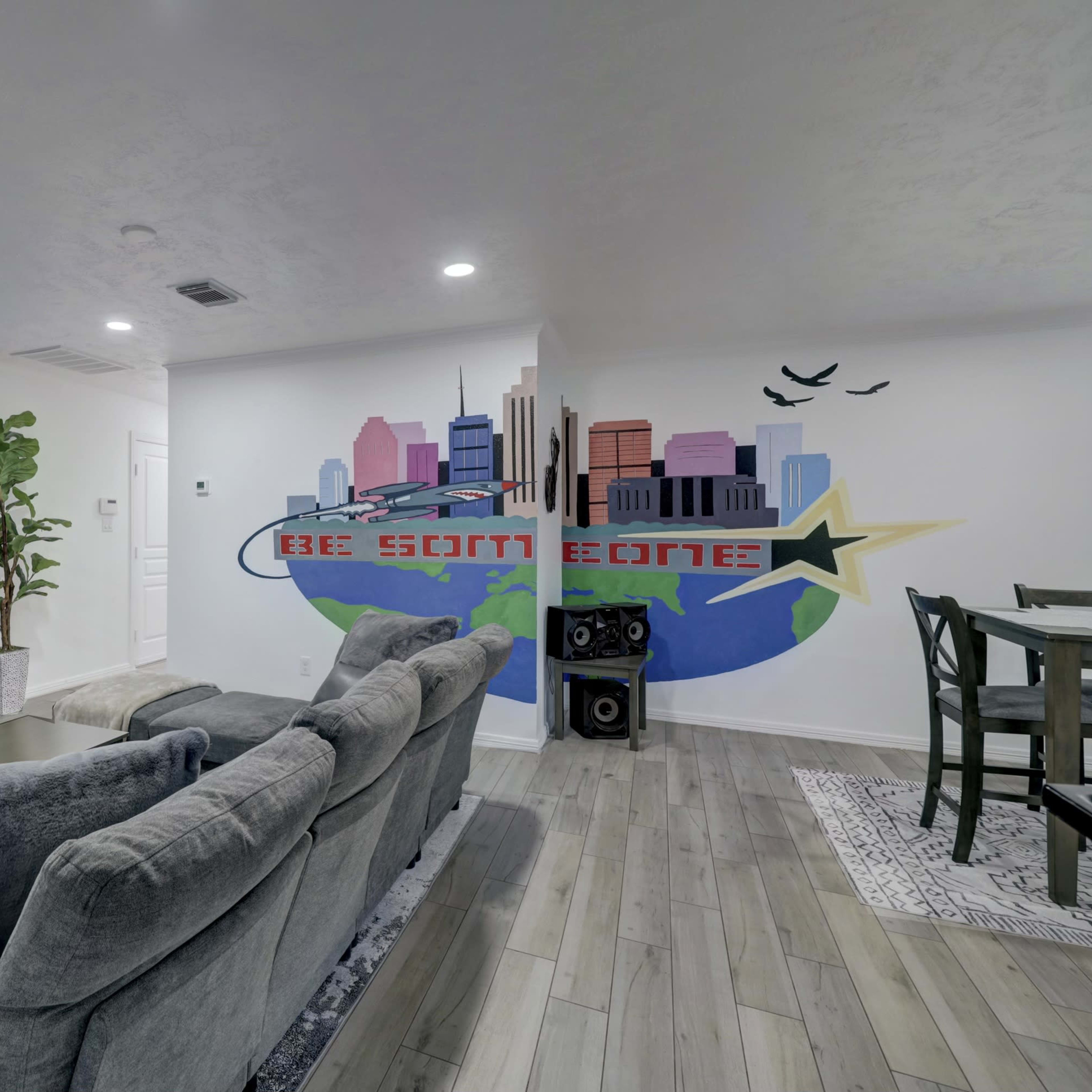 A modern living room features a sleek fireplace, a large TV mounted on the wall, and a mural depicting a city skyline with the phrase "BE SOMEONE" beside it.