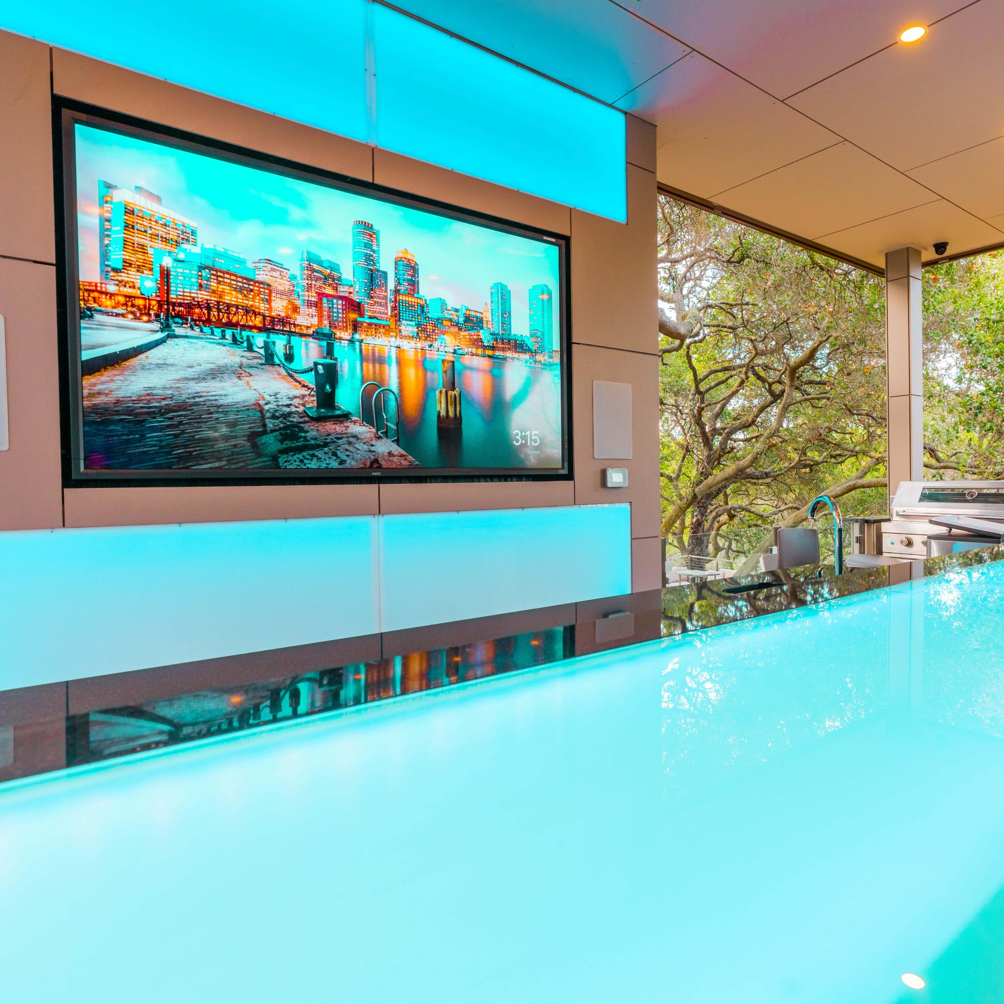 A modern outdoor bar area with a large screen displaying a cityscape, illuminated by blue ambient lighting.
