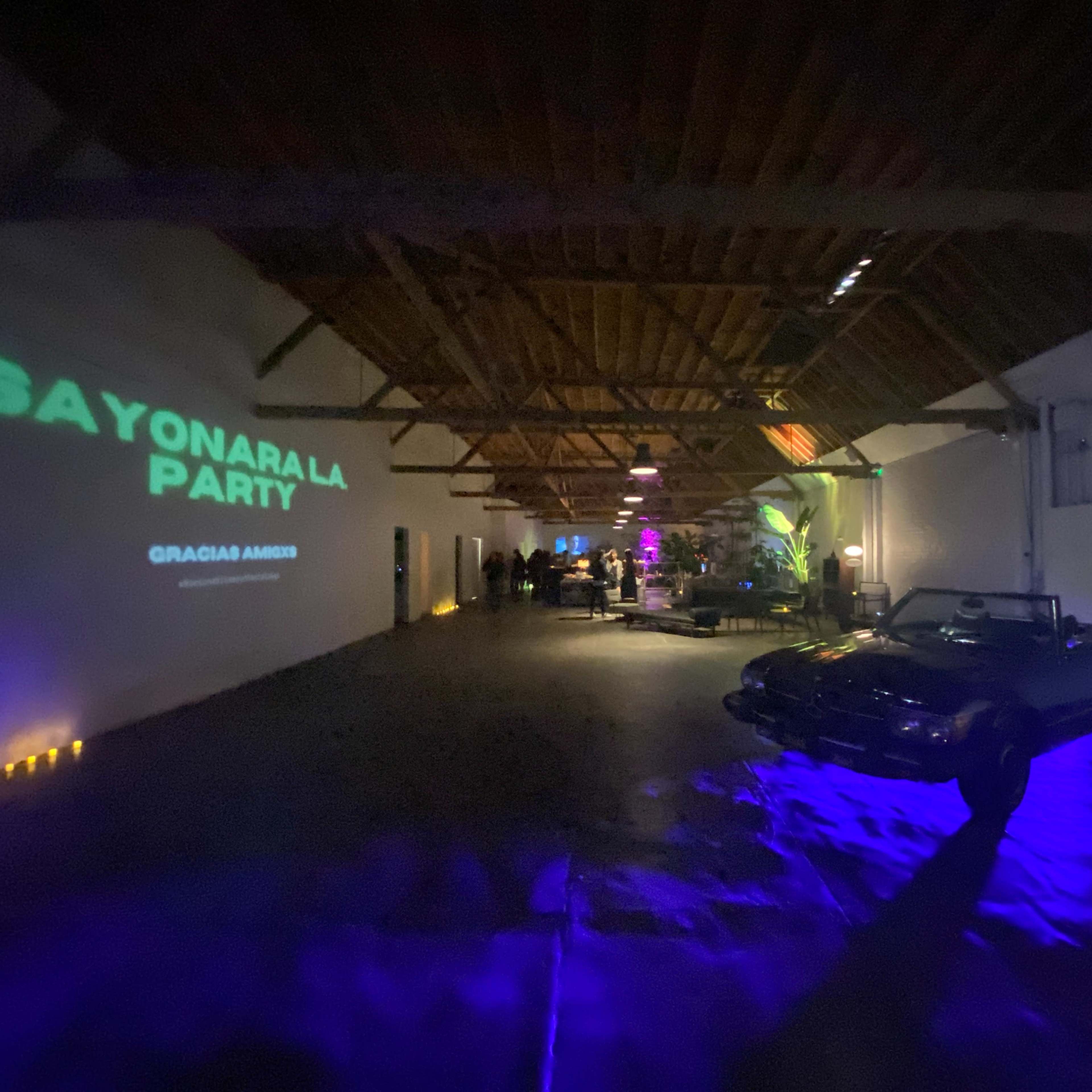 A spacious indoor venue features a classic convertible car, illuminated by colorful lights, while guests gather for an event titled "Sayonara La Party."