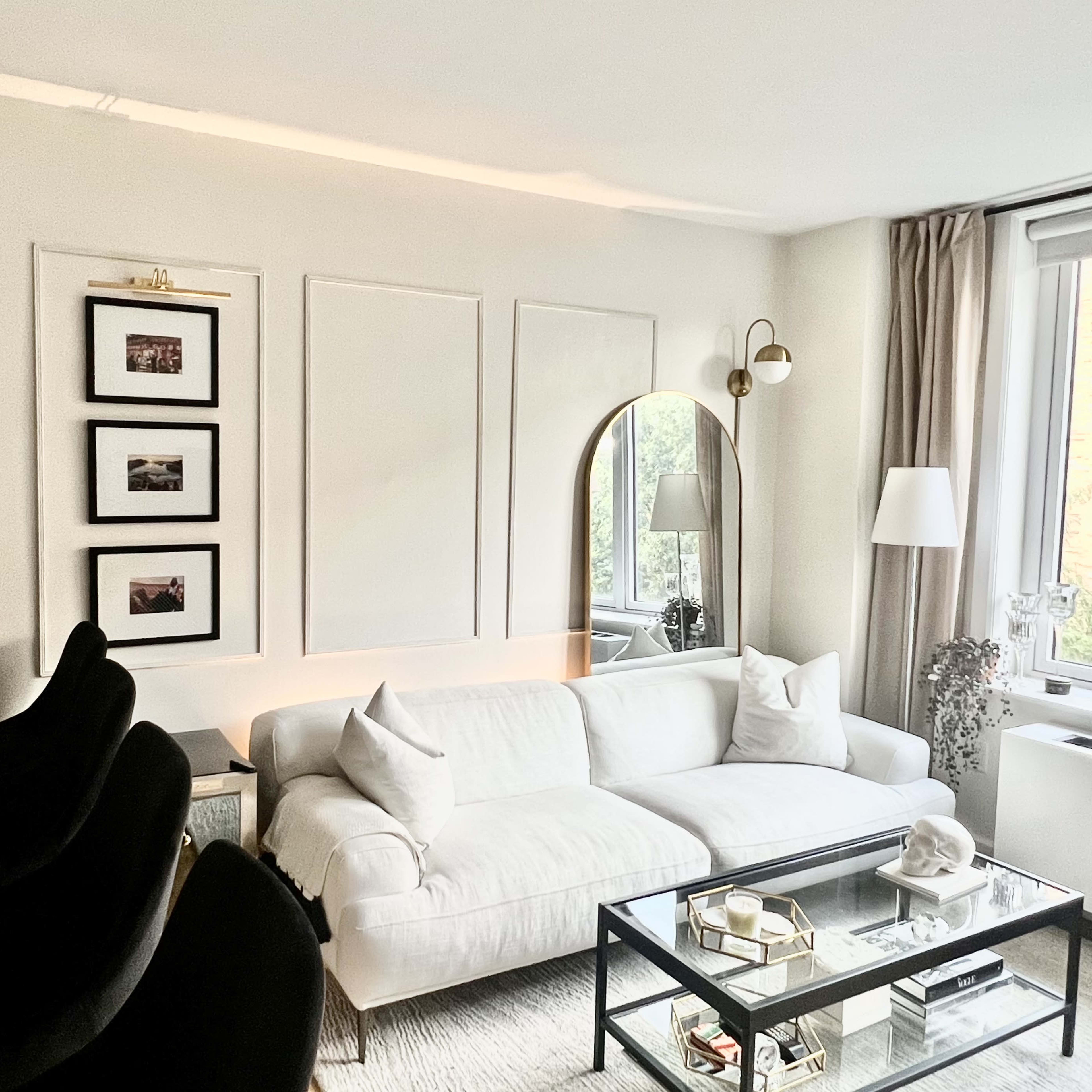 A modern living room features a white sofa, a glass coffee table, framed pictures on the wall, and large windows with a view of greenery outside.