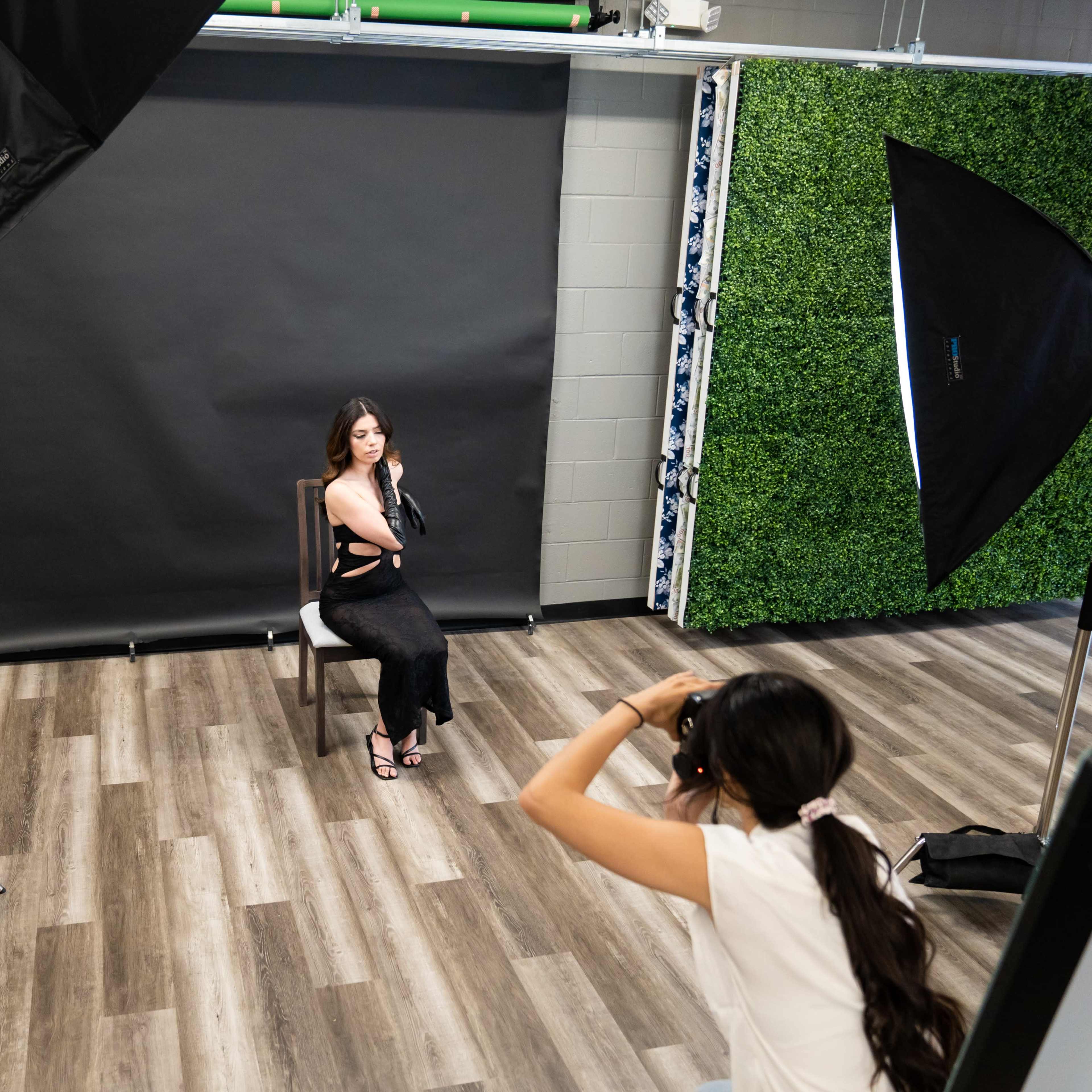 A woman poses in a chair for a photoshoot while another woman adjusts her camera settings.