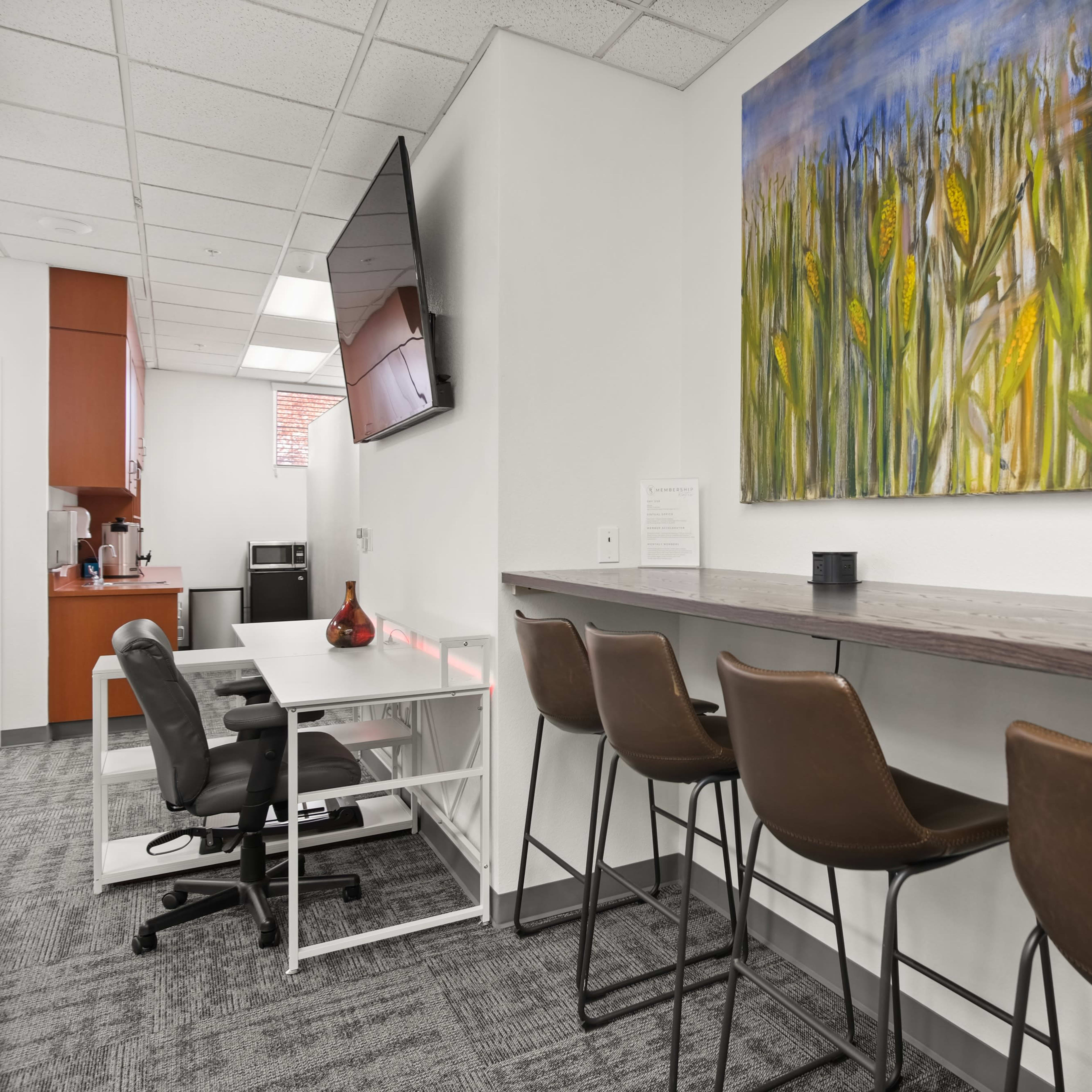 A modern workspace featuring a bar-style table with four brown stools, a black chair at a desk, and a colorful painting of cornfields on the wall.