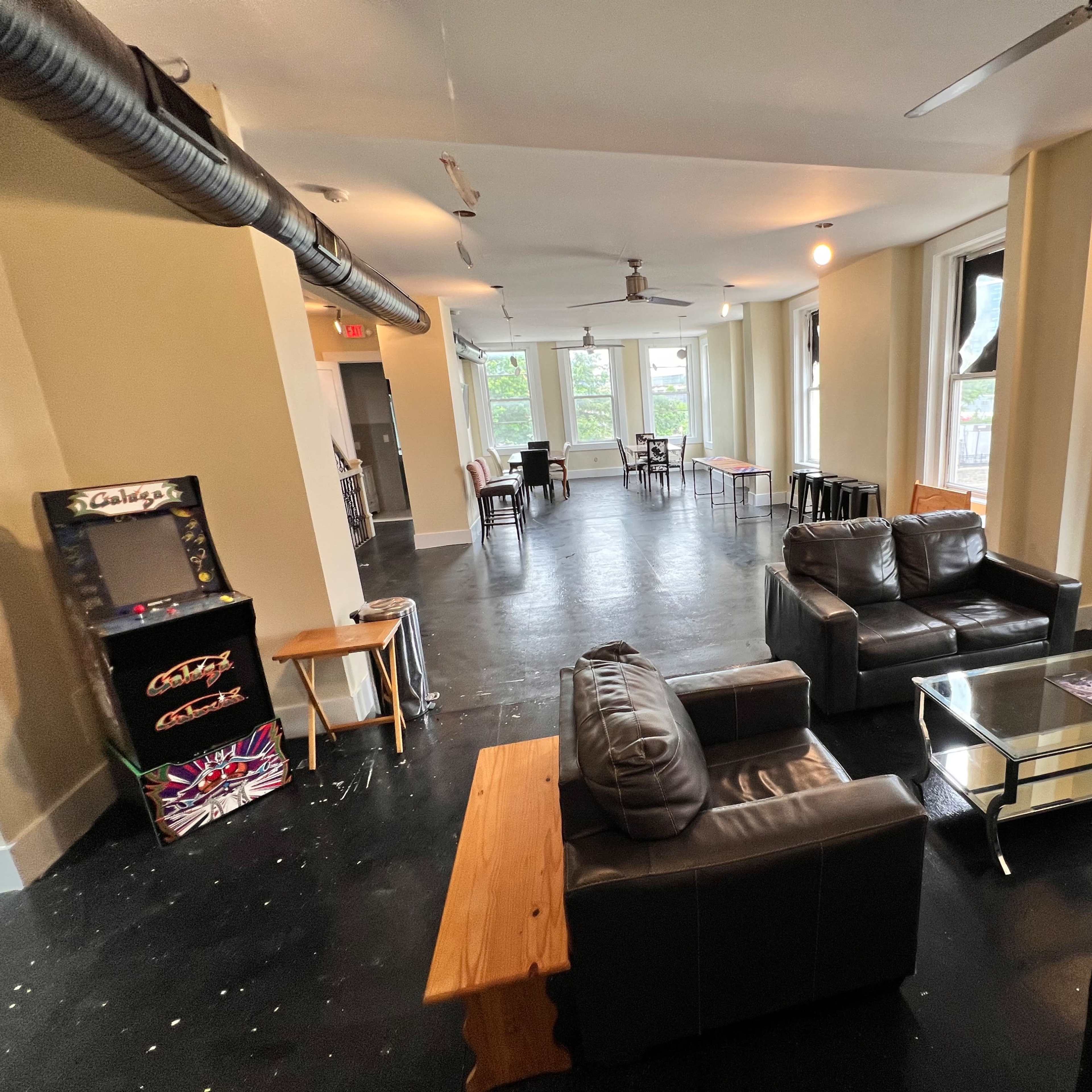 An open lounge area with black sofas, a glass coffee table, and several tables and chairs in the background, along with a vending machine in one corner.