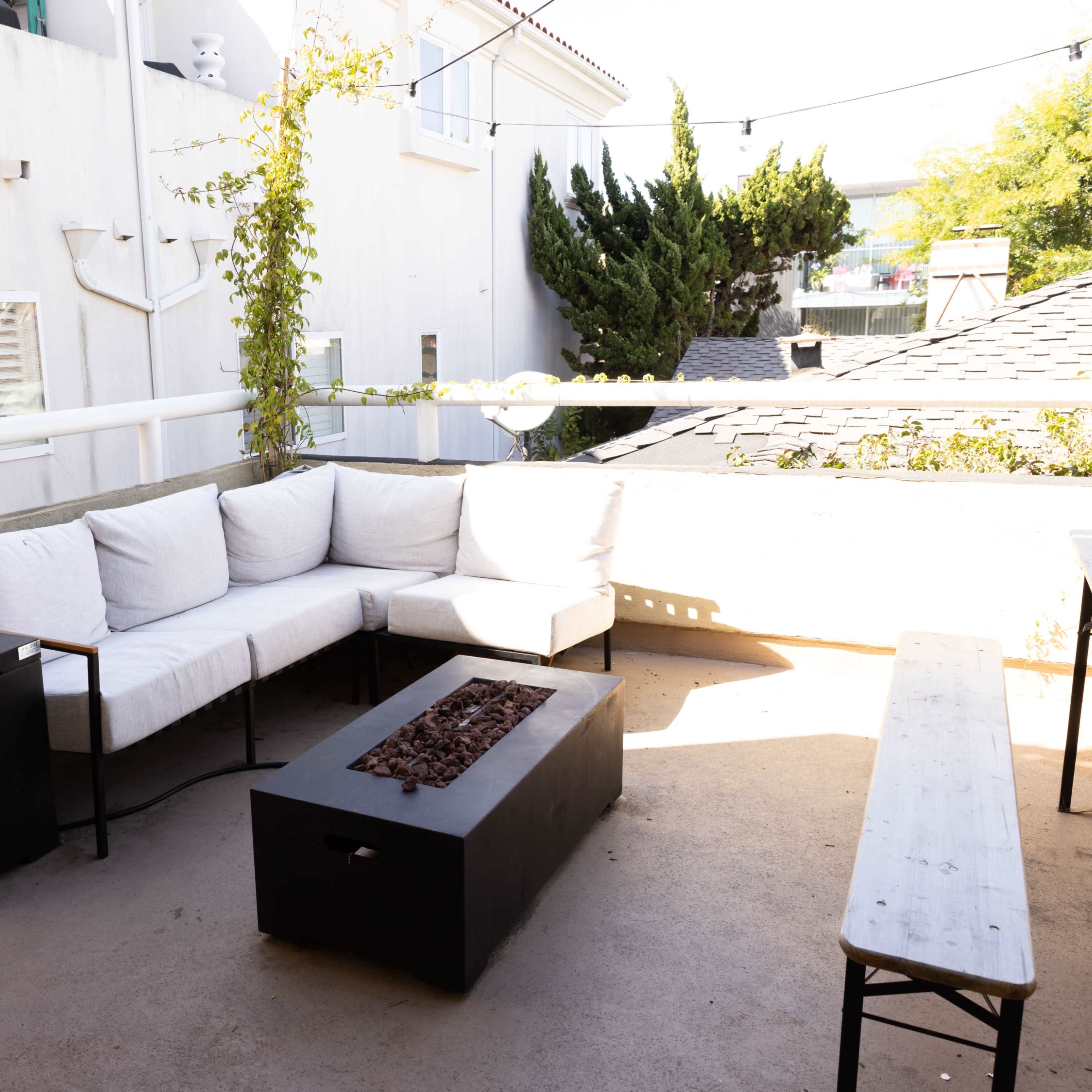 A small outdoor patio with a sectional sofa, a fire pit at the center, and a long wooden bench along one side.