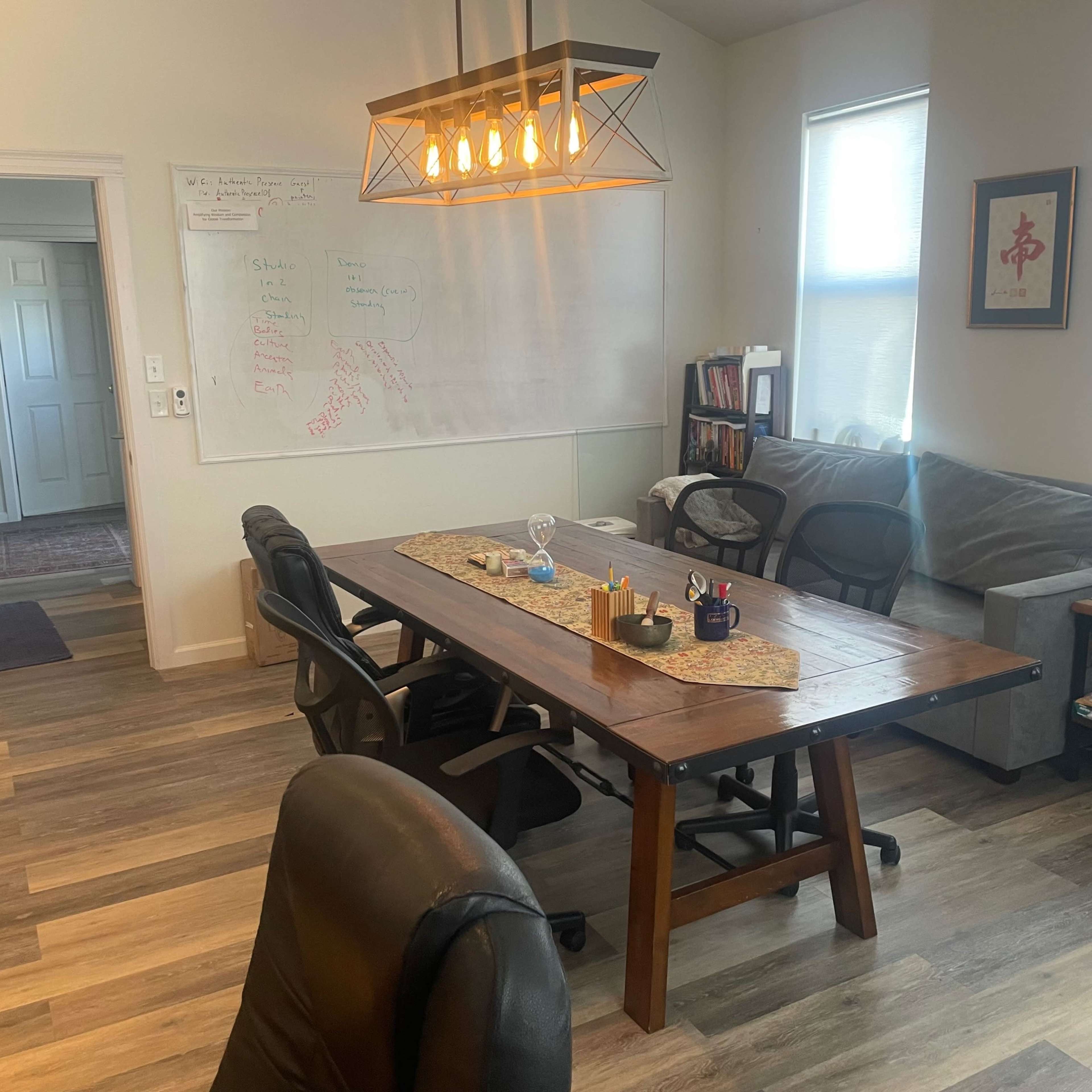 A brightly lit office space featuring a large wooden table with chairs, a whiteboard with notes, and bookshelves along the wall.