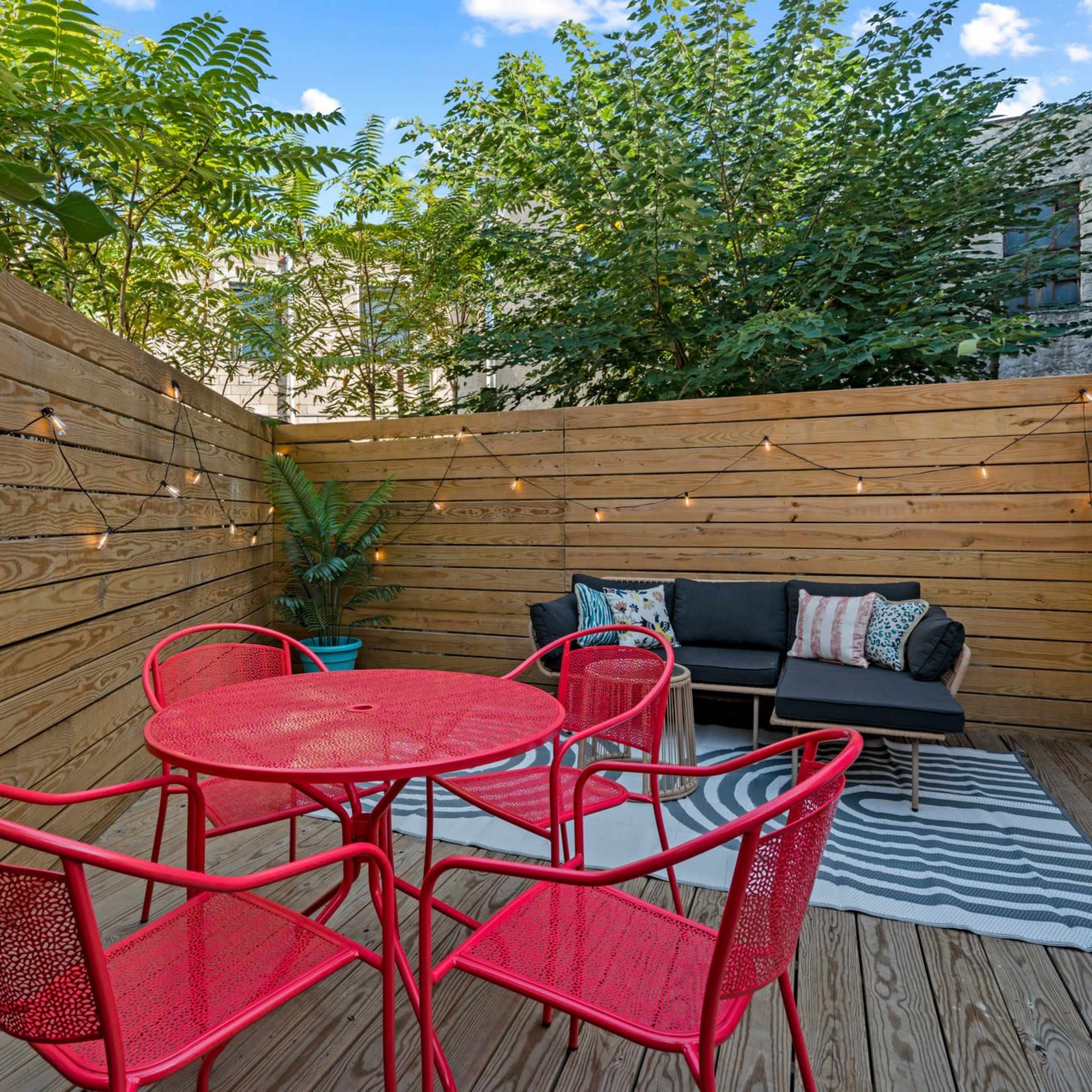 A small outdoor seating area with a red metal table and chairs, a black sofa, and string lights, surrounded by wooden fences and greenery.