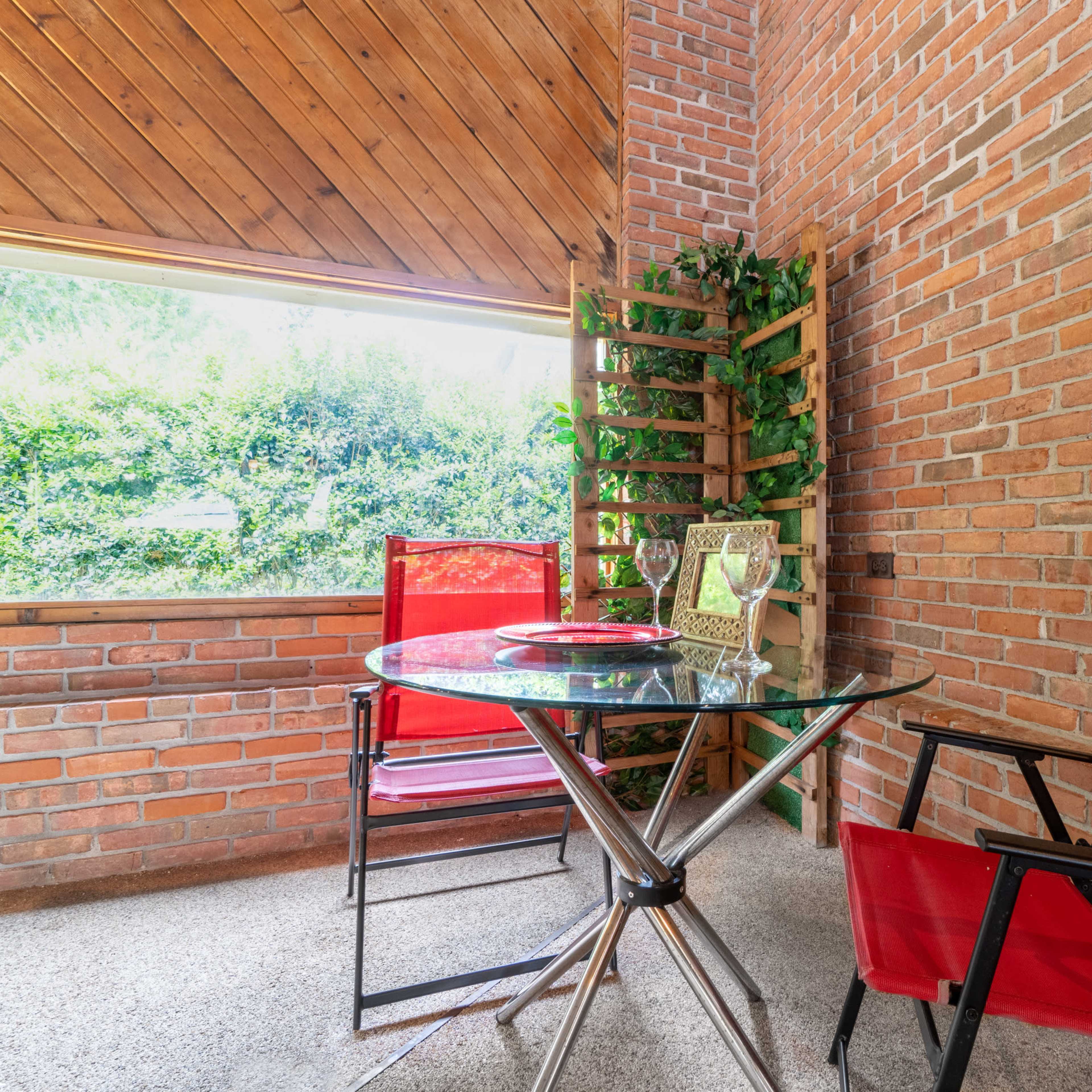A small terrace features a glass table and two red chairs beside a wooden trellis with greenery.