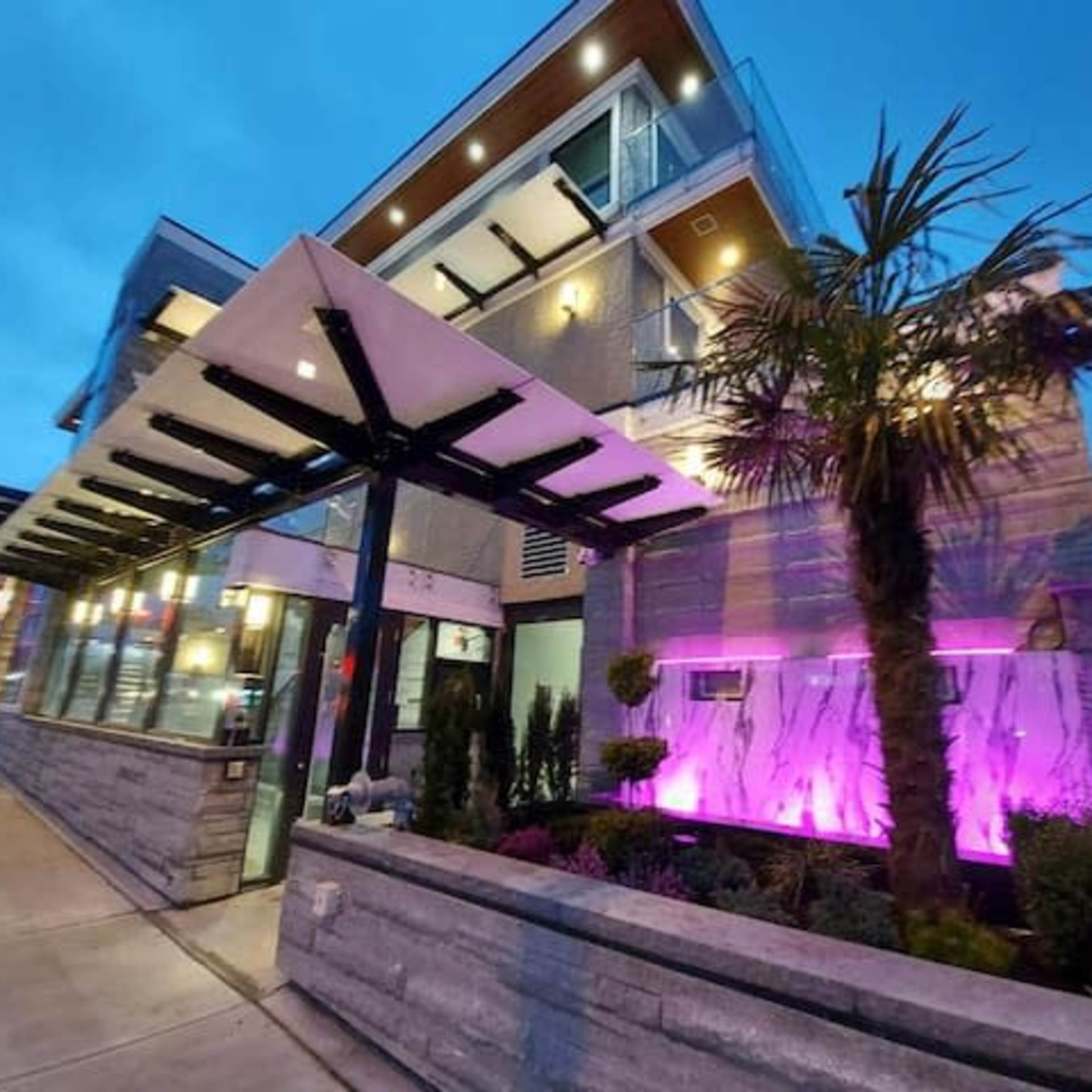 A modern building with a flat roof, featuring large windows and illuminated landscaping with palm trees, set against a twilight sky.