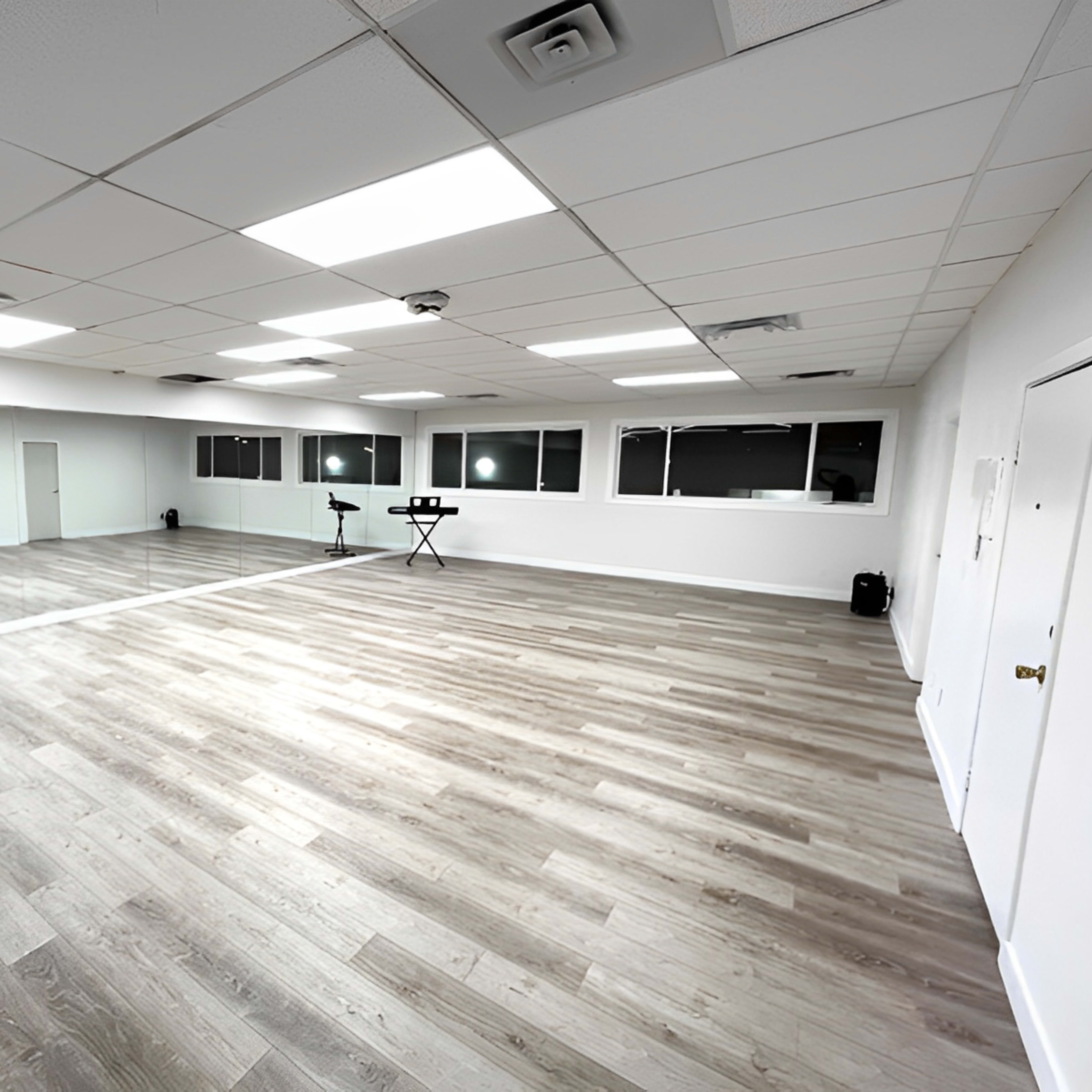 An empty dance studio features a large wooden floor, mirrors on one wall, and bright overhead lighting.