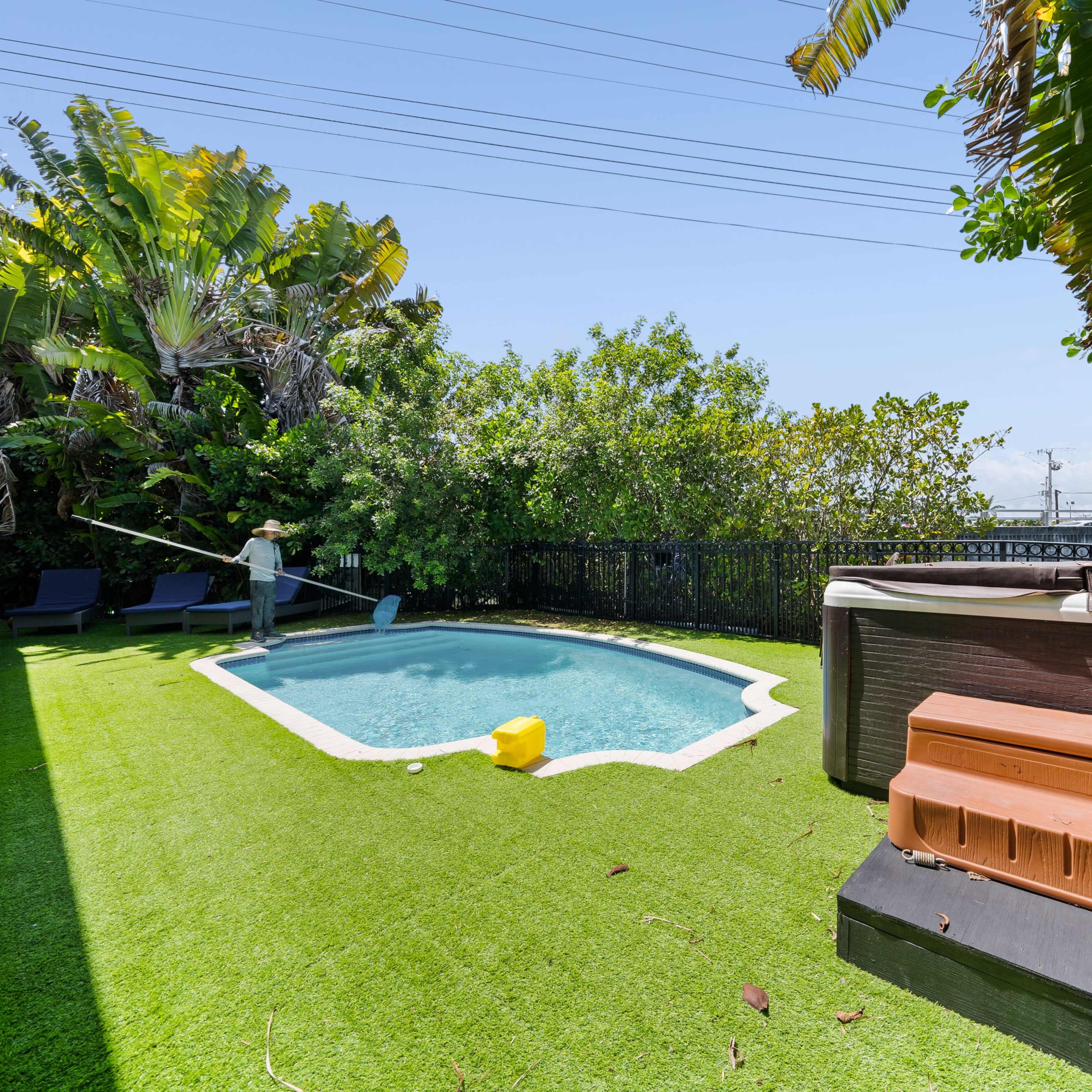 A backyard with a pool, a hot tub, artificial grass, and lush greenery surrounding the area.