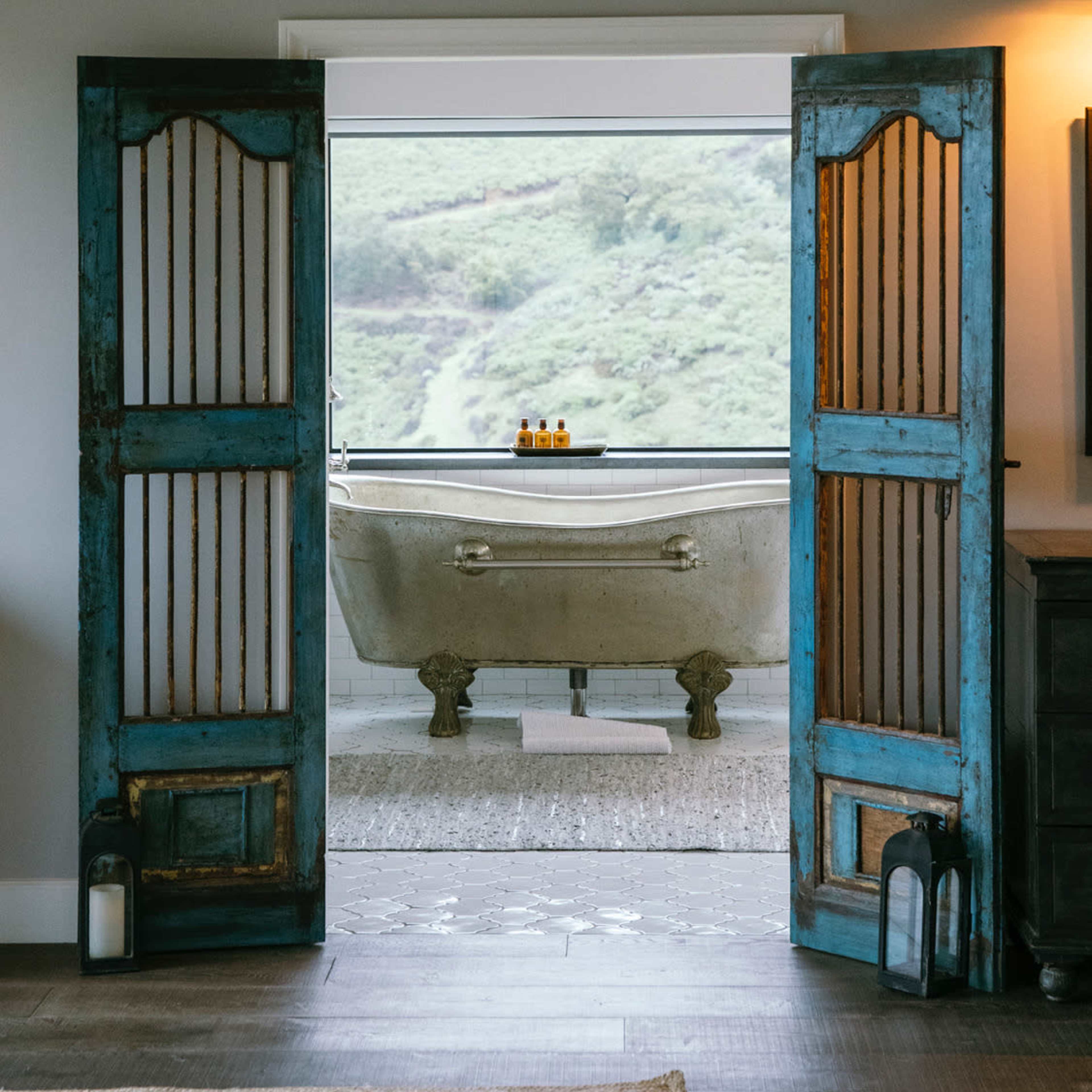 An antique bathtub is positioned in front of an open window, framed by blue wooden doors, revealing a scenic view of green hills.