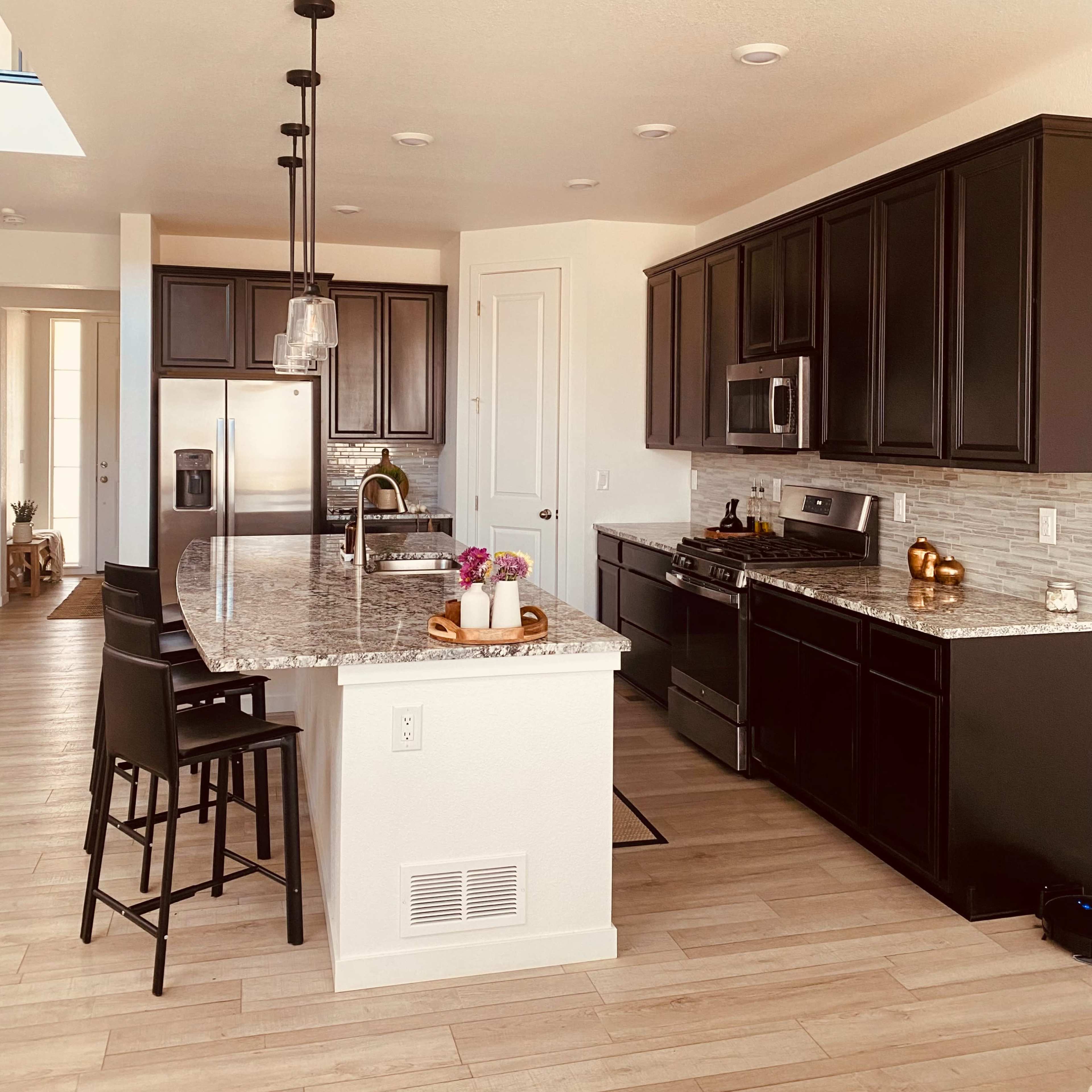 A modern kitchen with dark cabinets, a granite countertop, and stainless steel appliances, featuring a large island with bar stools.