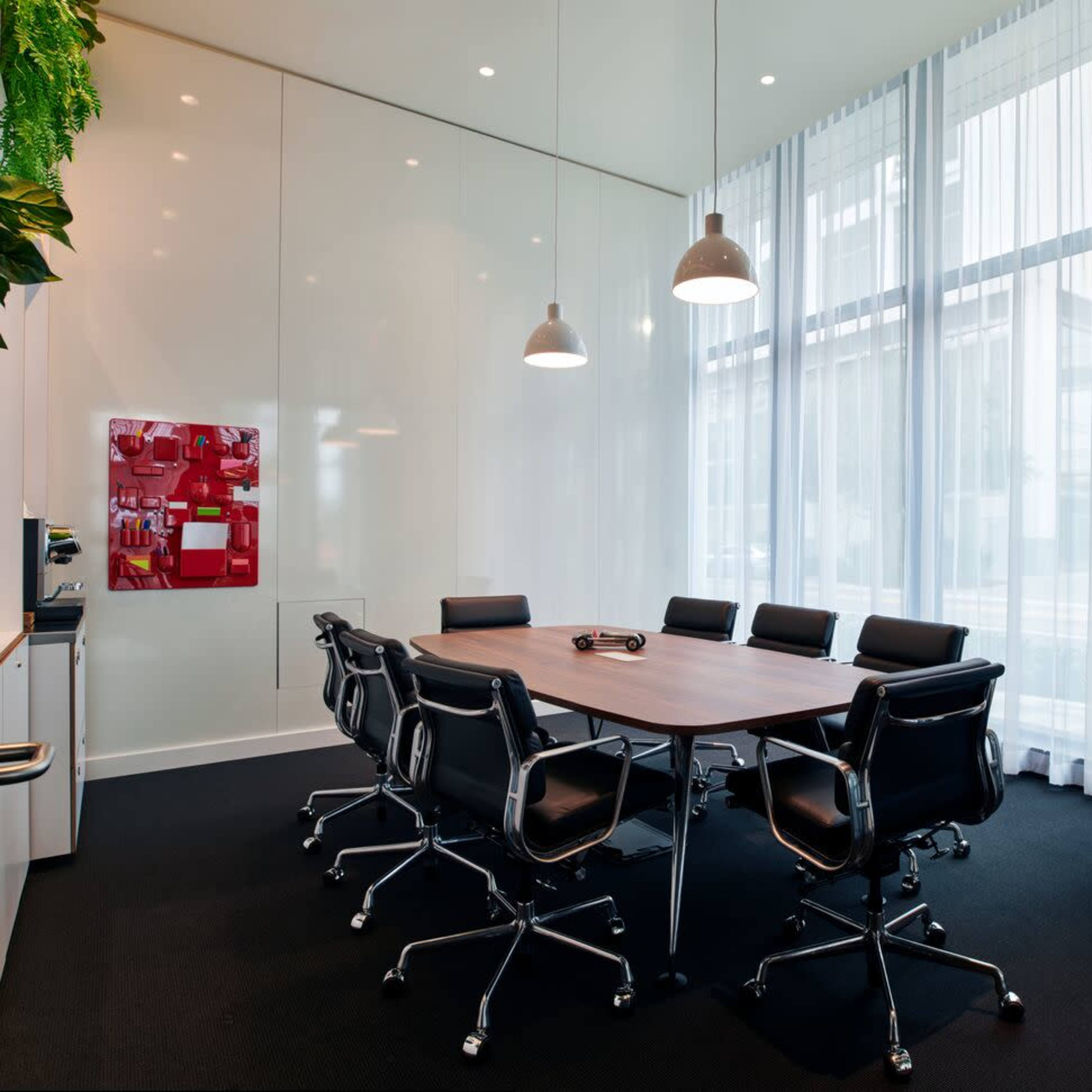 A conference room with a large wooden table surrounded by black leather chairs, illuminated by pendant lights, and featuring floor-to-ceiling curtains.