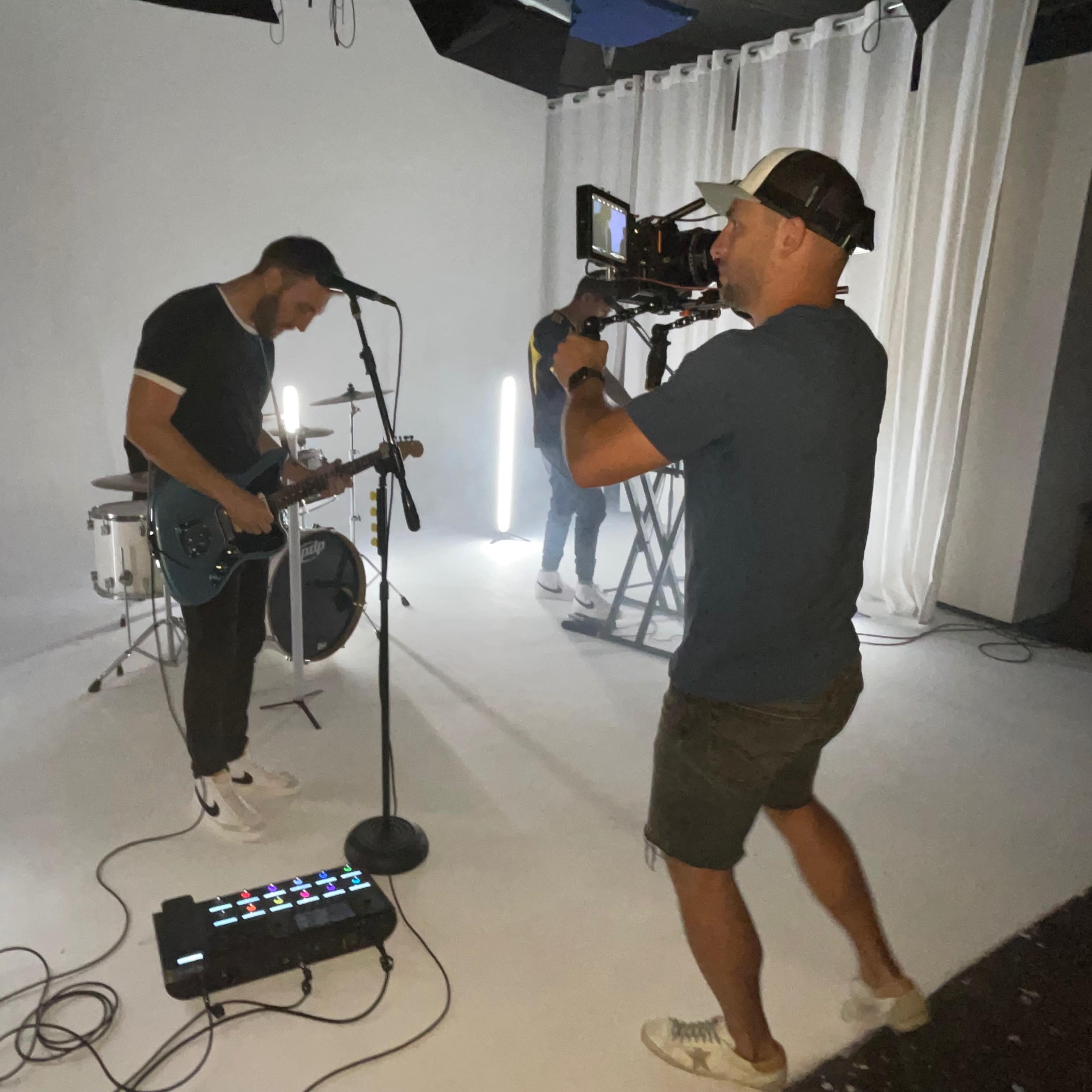 A musician plays guitar and sings on a white backdrop while a camera operator films him.