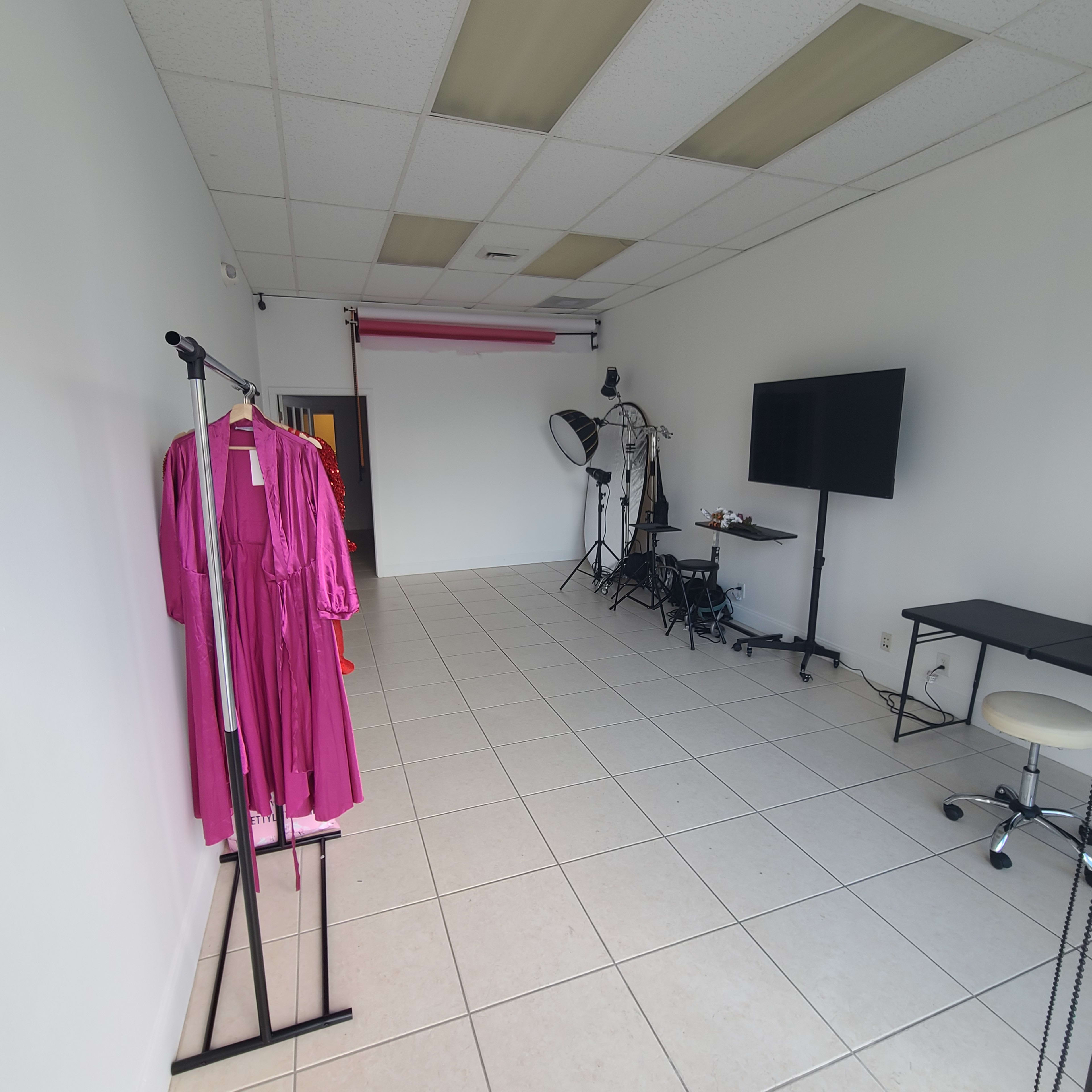 A spacious studio with white walls, tiled flooring, and various photography equipment, including a large monitor, a light setup, and a rack holding a pink dress.