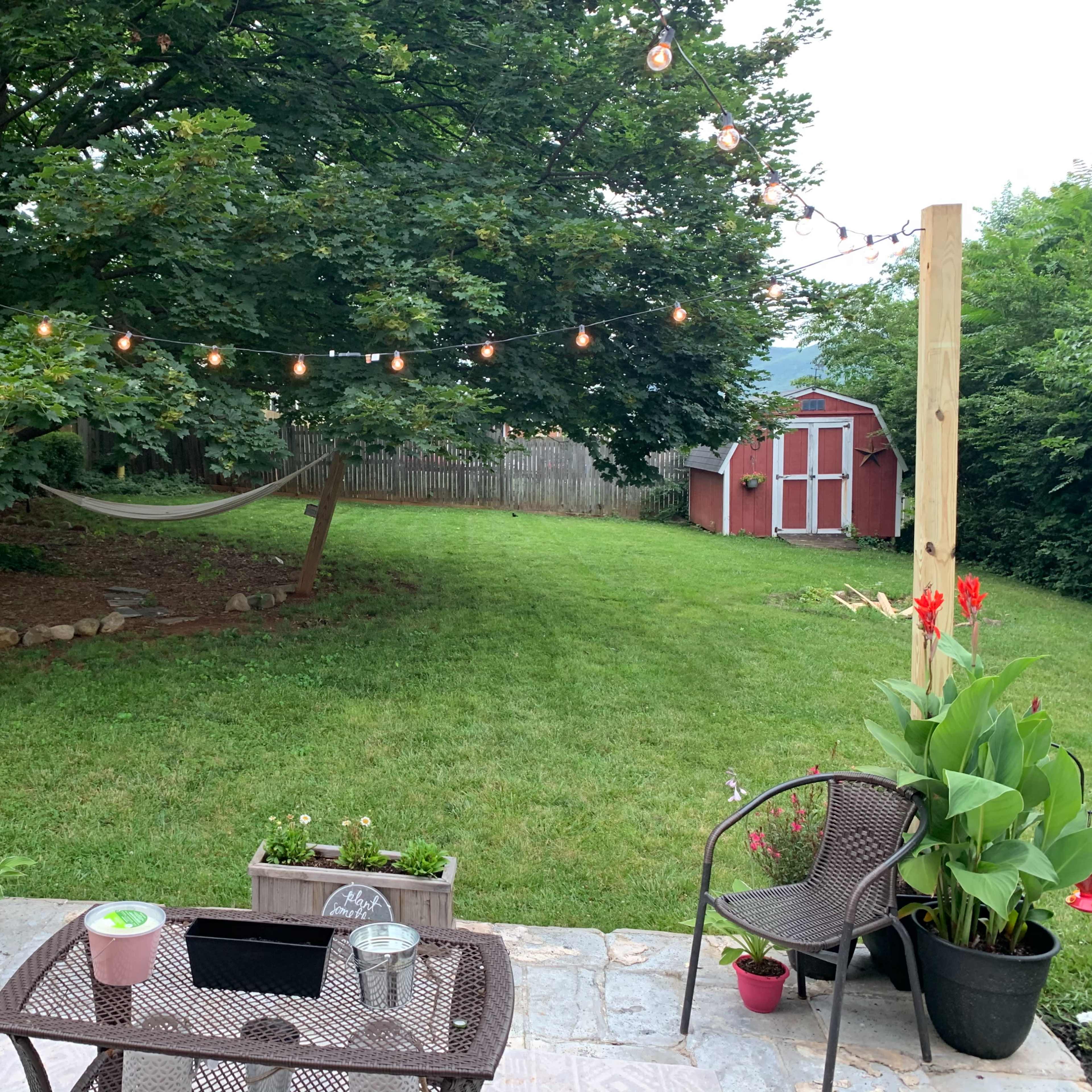 A backyard scene features a hammock strung between two wooden posts adorned with string lights, flanked by potted plants and two chairs on a stone patio.