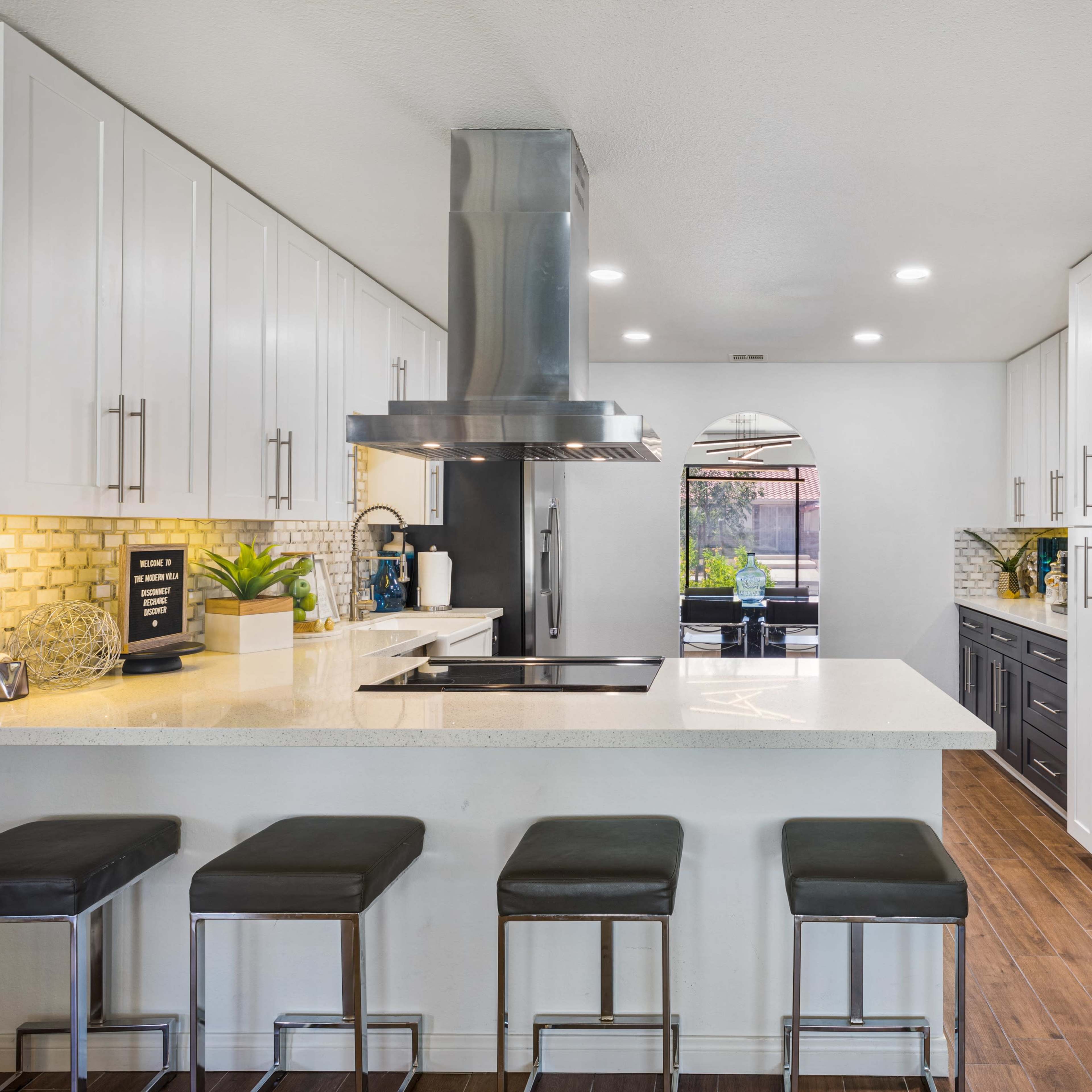 A modern kitchen features a central island with bar stools, stainless steel appliances, and dark cabinetry against light walls.