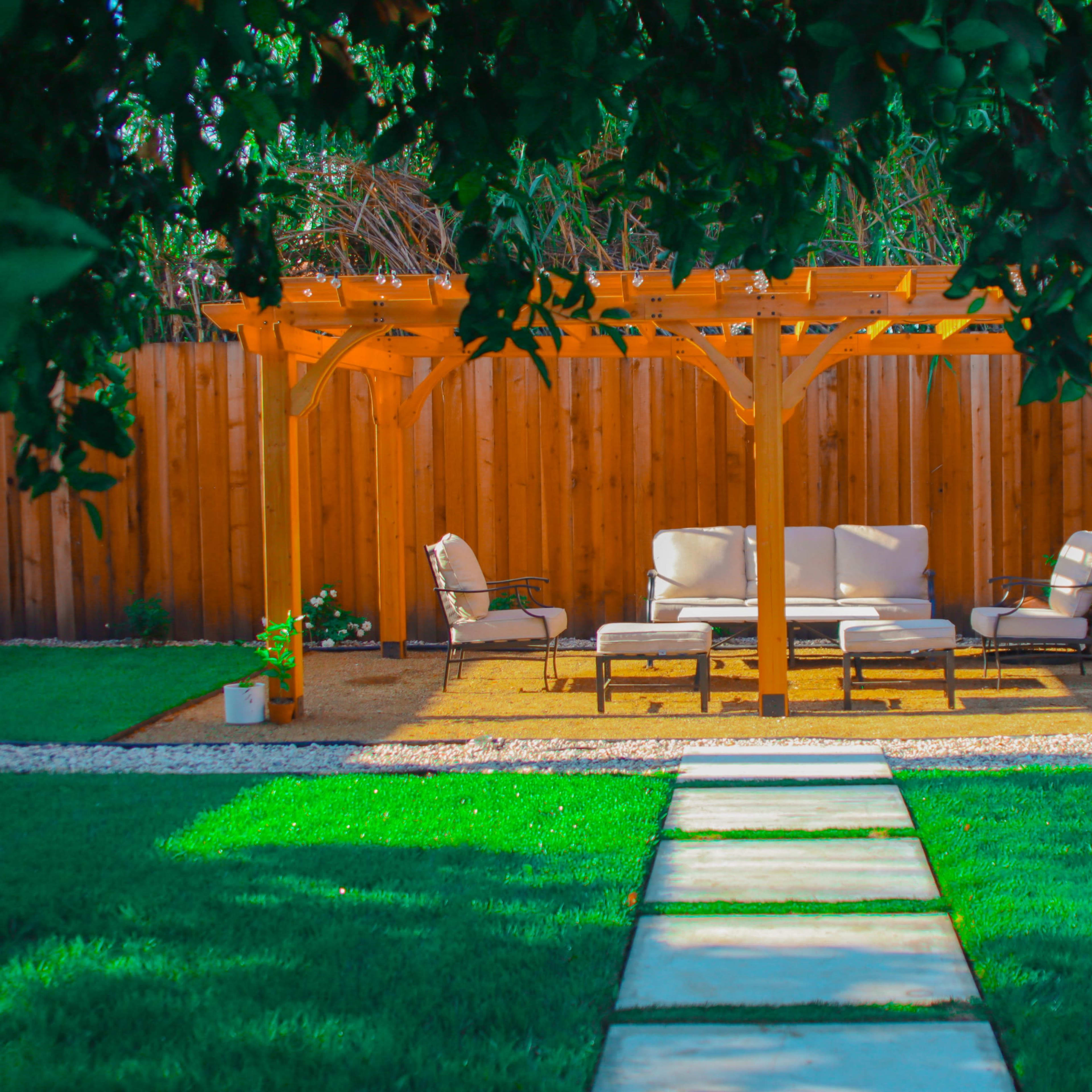 A wooden pergola provides shade over a set of beige outdoor furniture in a landscaped backyard featuring a stone pathway and well-manicured grass.