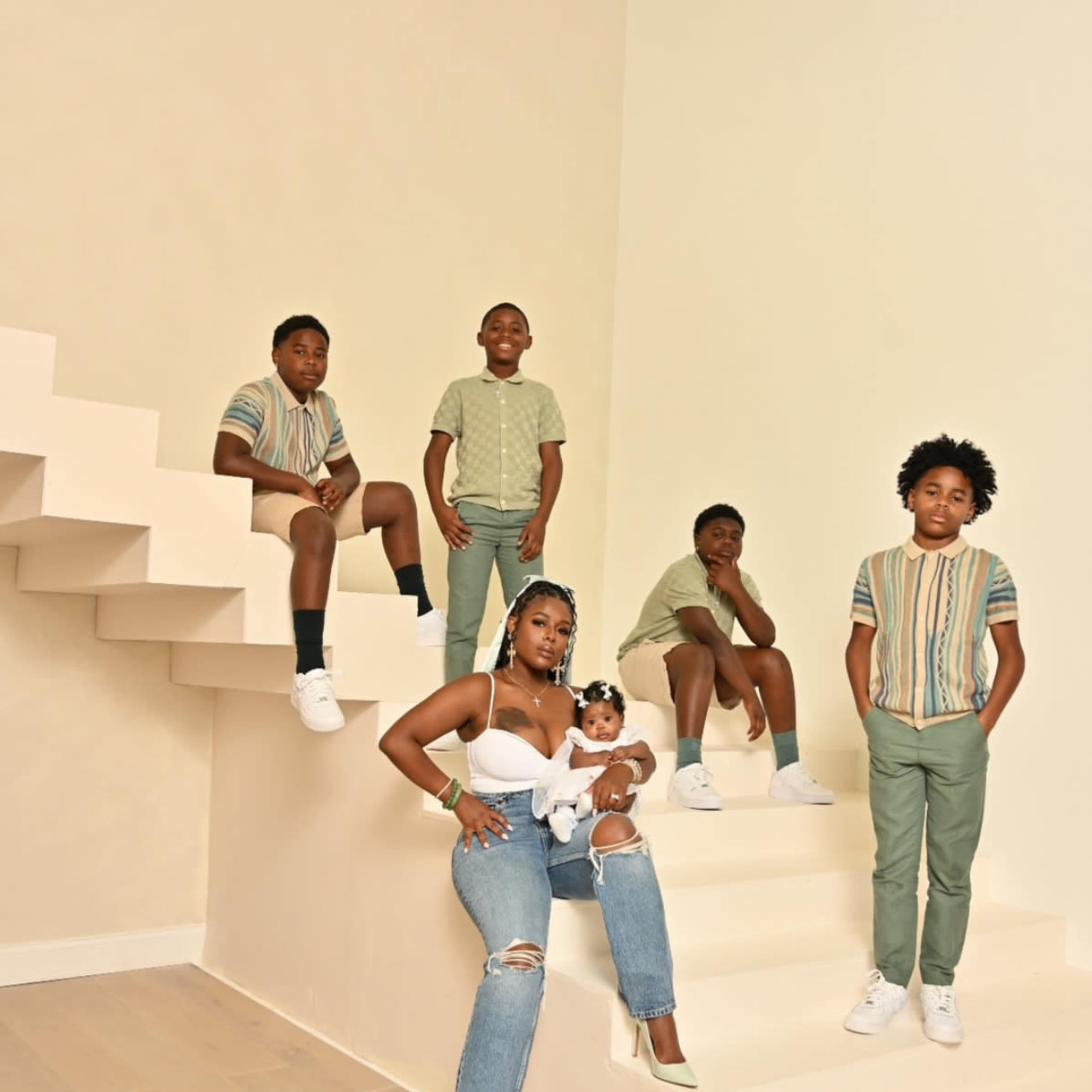 A woman sits on a staircase holding a baby, while four children stand or sit around her in casual outfits.