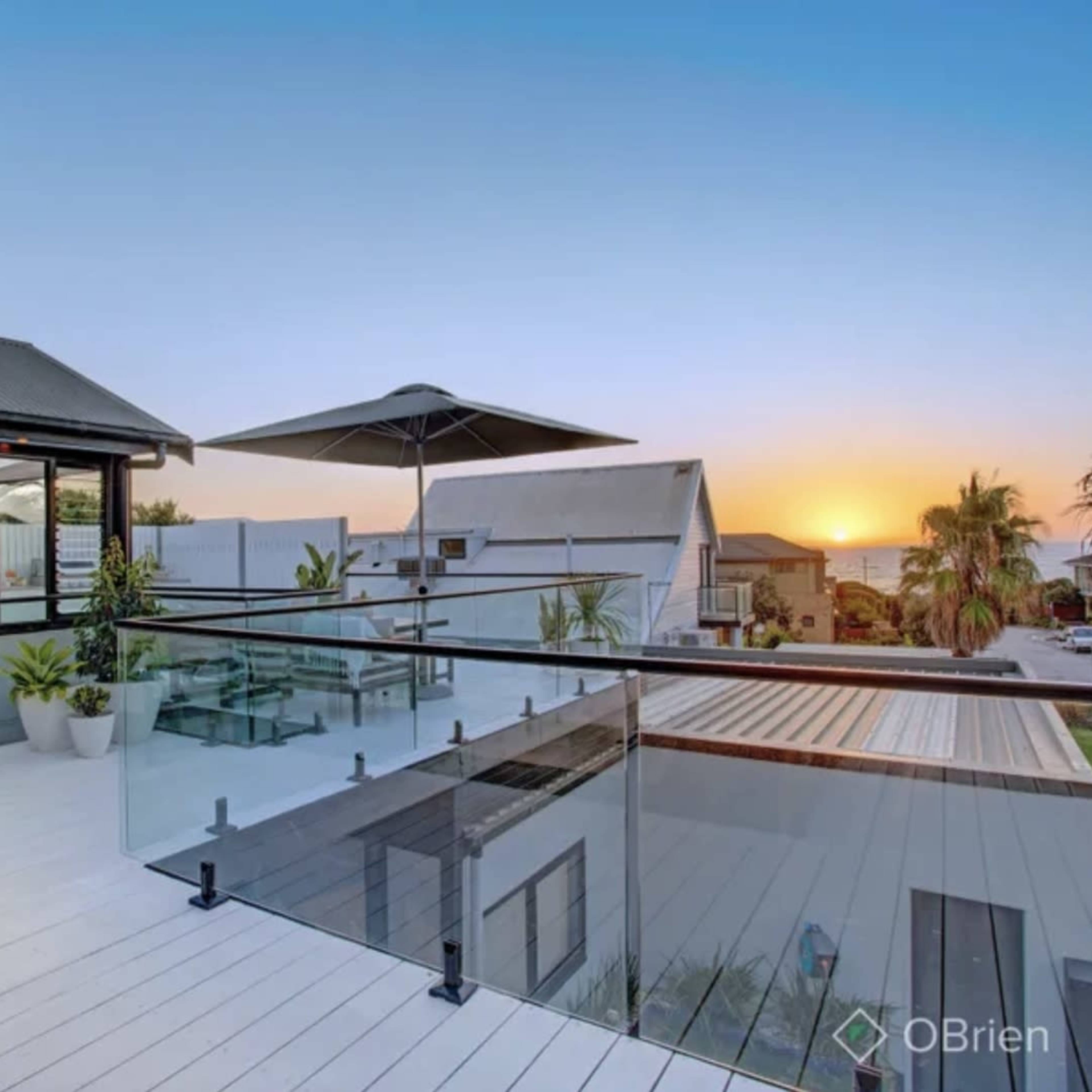 A modern balcony with a glass railing overlooking a sunset on the horizon, surrounded by palm trees and residential buildings.