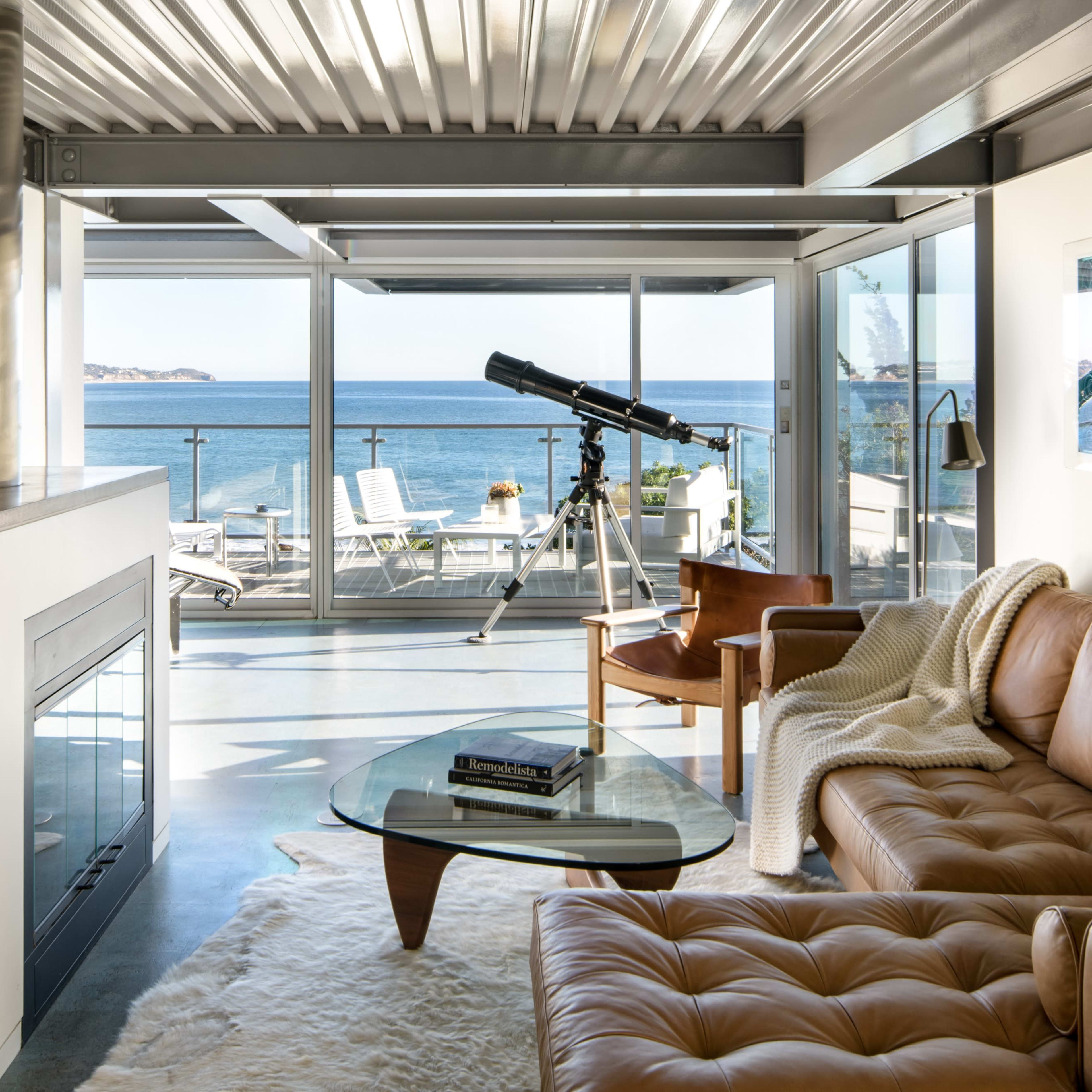 The living room features a leather sofa, a glass coffee table, and a telescope, all overlooking a scenic ocean view through large glass windows.