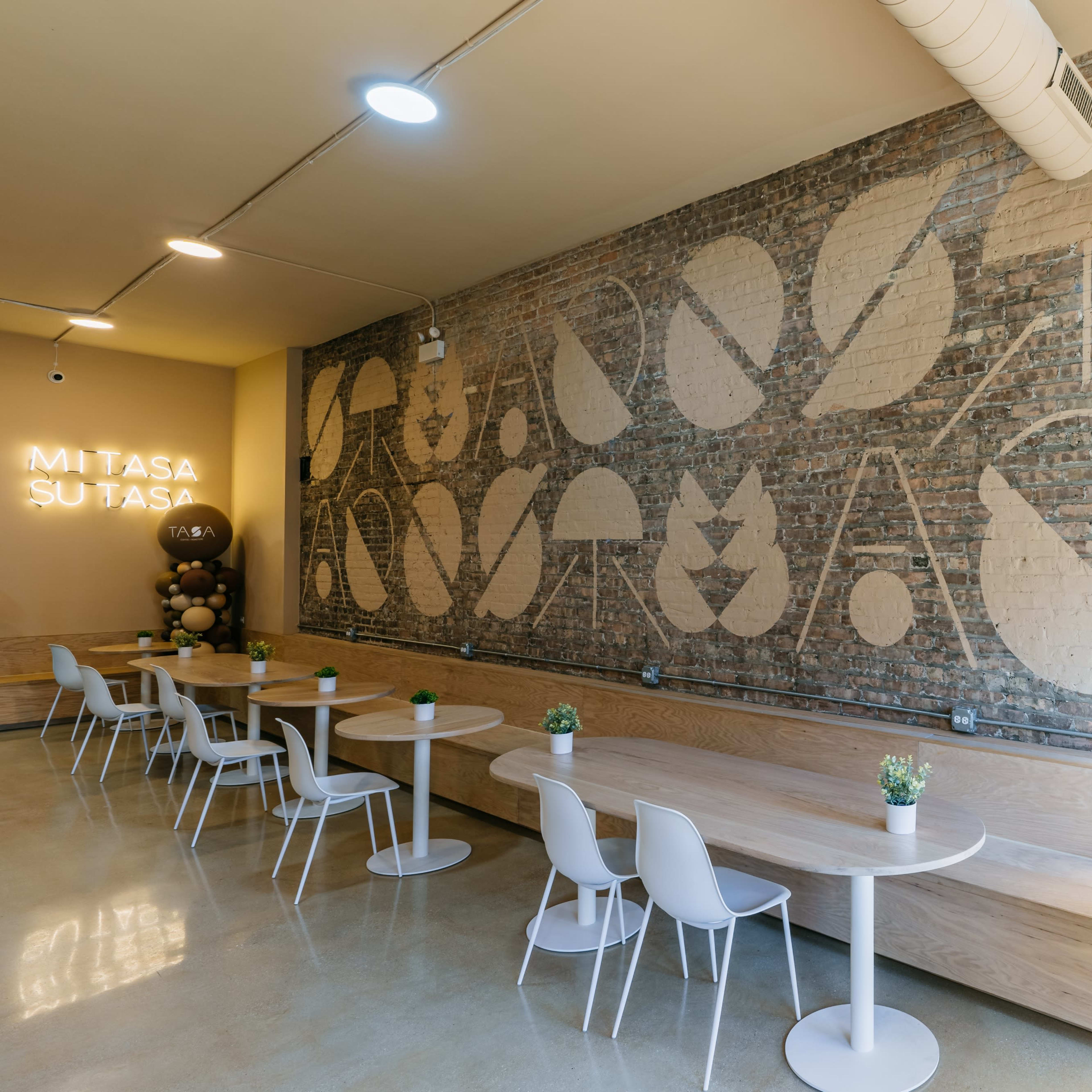 A modern interior with wooden tables and white chairs, set against a brick wall featuring a large abstract mural and illuminated signage.
