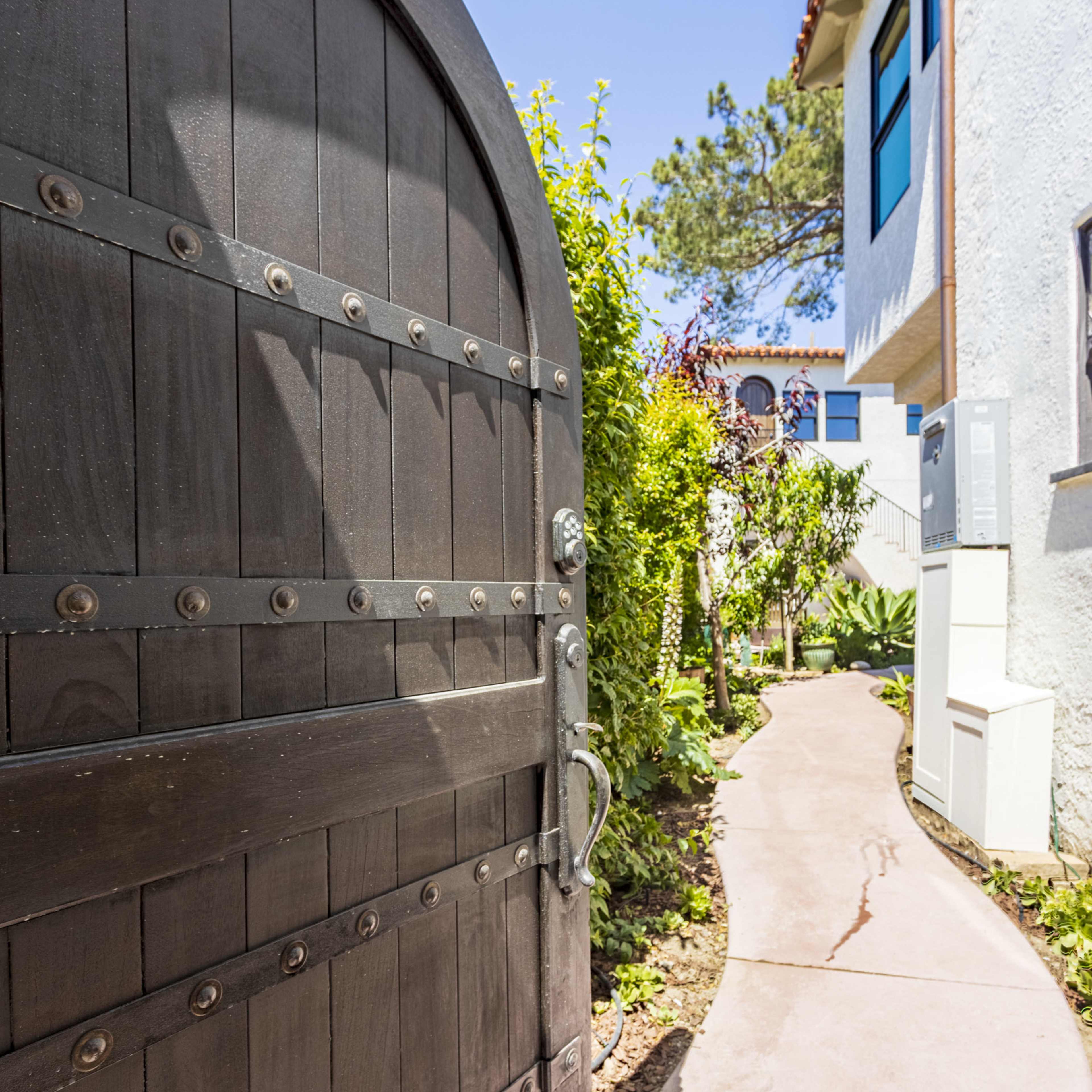 A black wooden gate stands open, revealing a landscaped pathway leading between a white building and greenery.