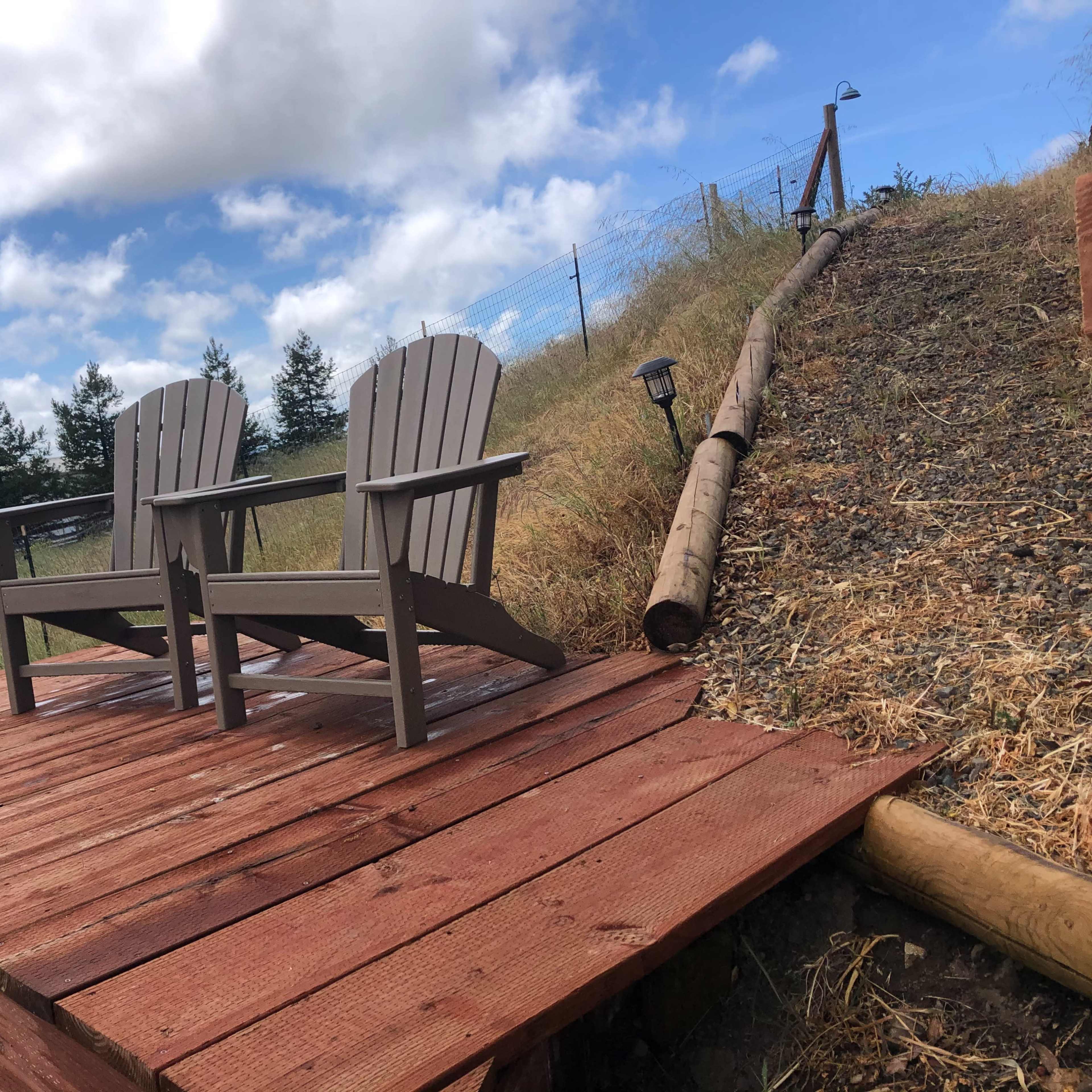 Two Adirondack chairs sit on a wooden deck overlooking a grassy slope with trees and a cloudy sky in the background.