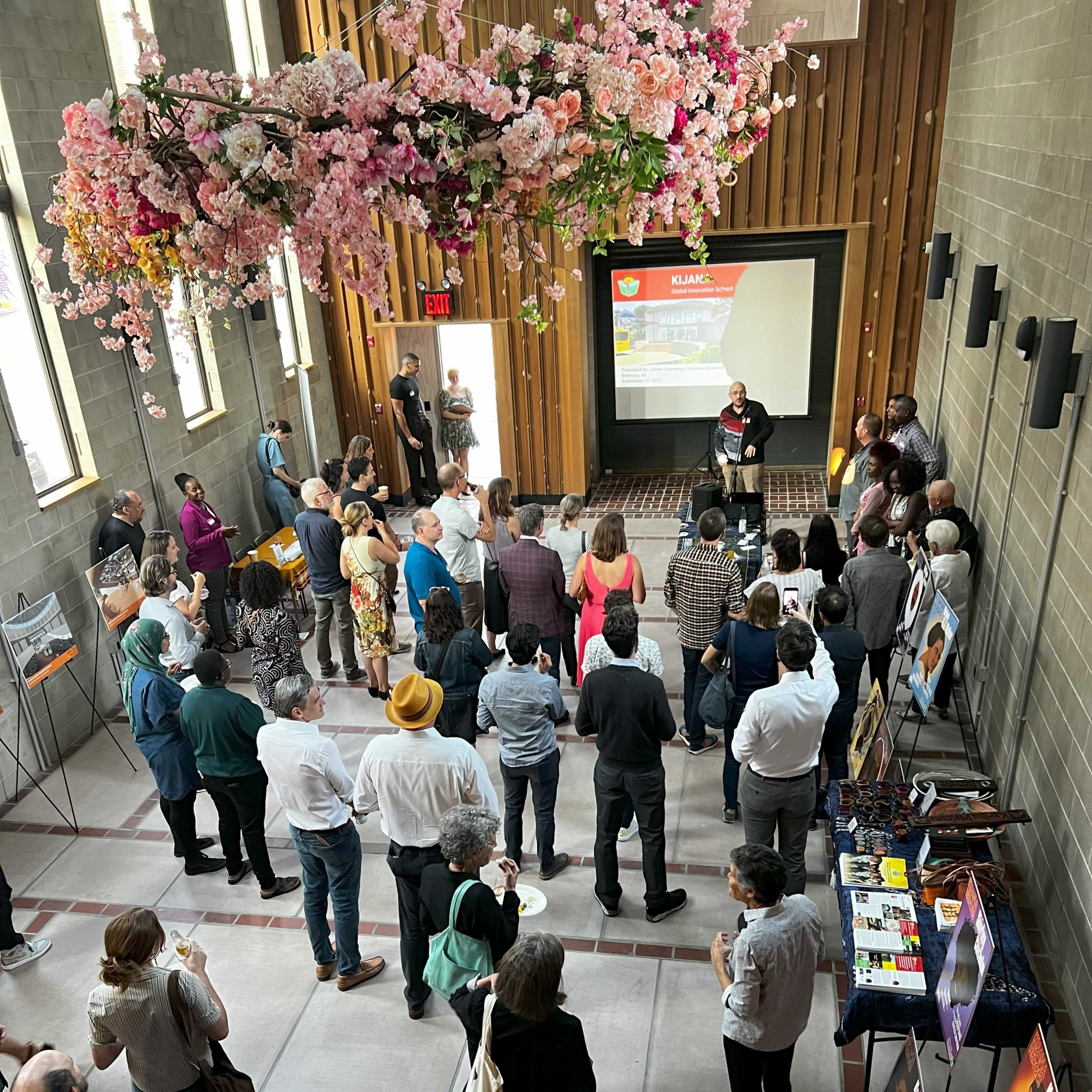 A crowd gathers in a room with wooden walls and a floral installation overhead, while a speaker presents in front of a projector screen.