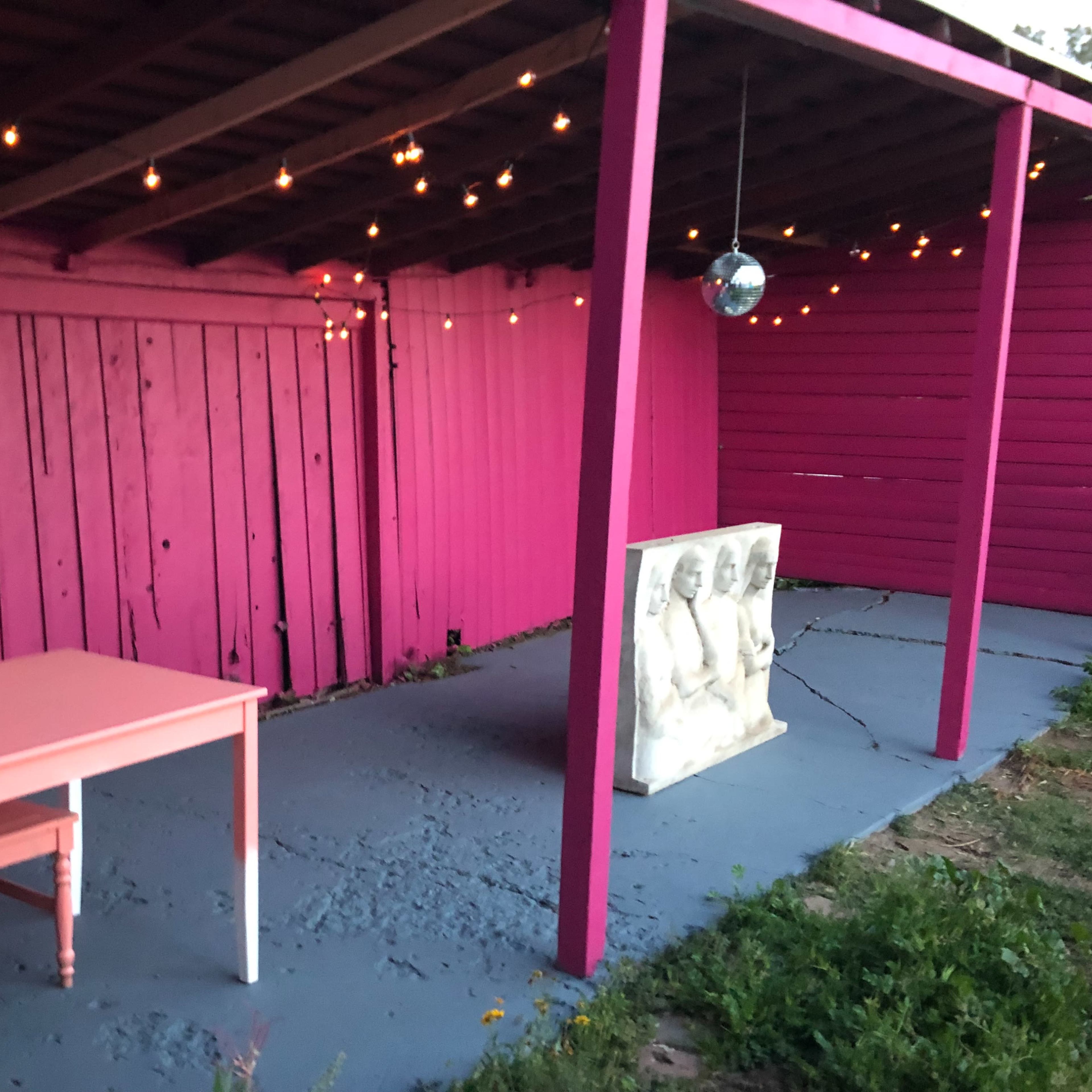 A small outdoor space featuring a pink wooden wall, a white table with matching chairs, and a sculpture of three figures, all illuminated by string lights overhead.